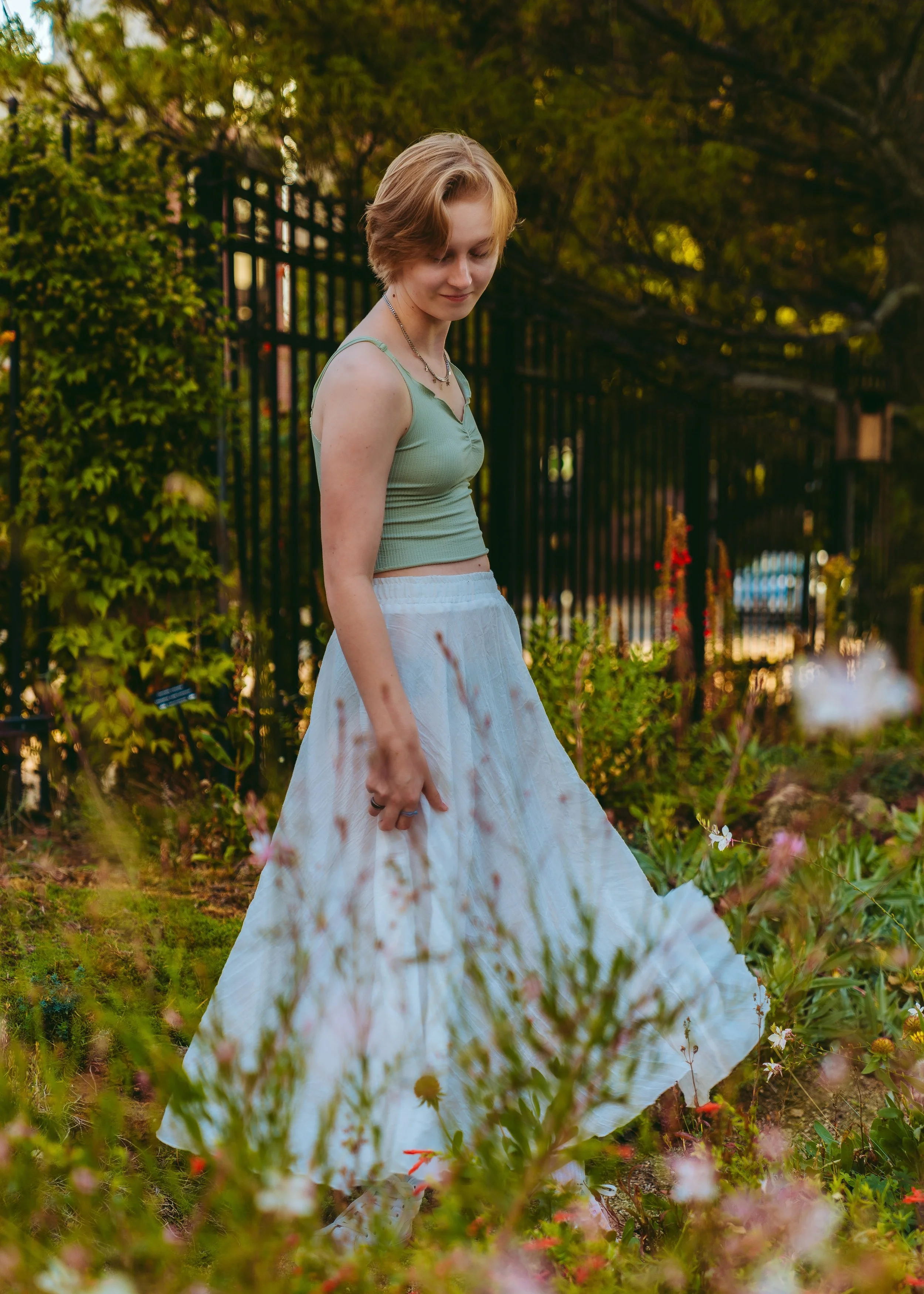 A young woman with short blonde hair wearing a light green tank top and a long white skirt, standing in a garden with flowers and greenery, over a black metal fence in the background, during late afternoon or early evening.