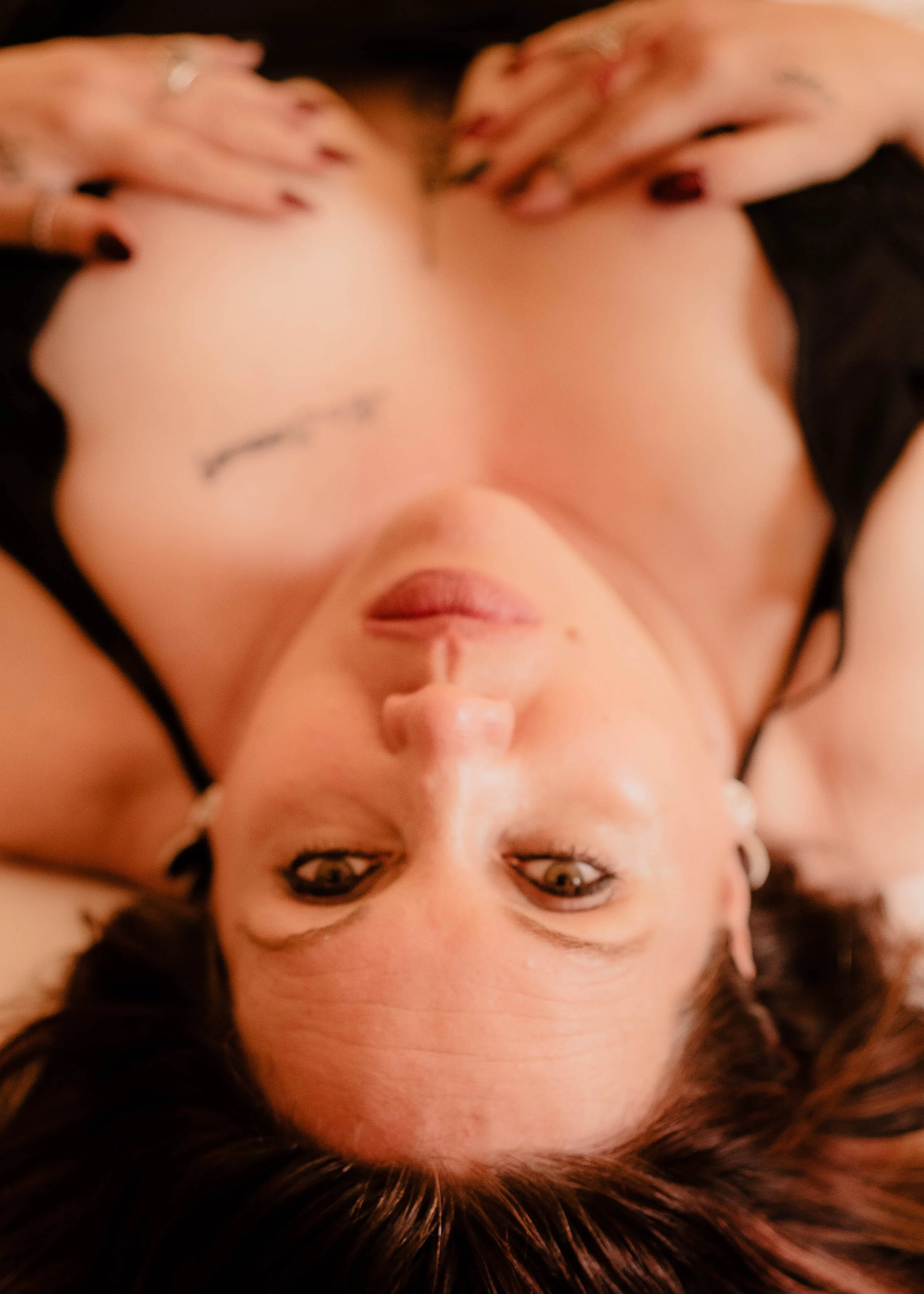 A woman lying down with her head upside down, looking directly at the camera, with dark hair and a tattoo on her chest.