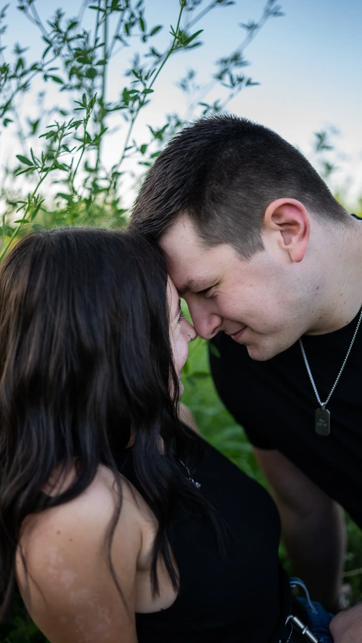 A couple with their foreheads touching, eyes closed, in an outdoor natural setting with green plants and a blue sky.