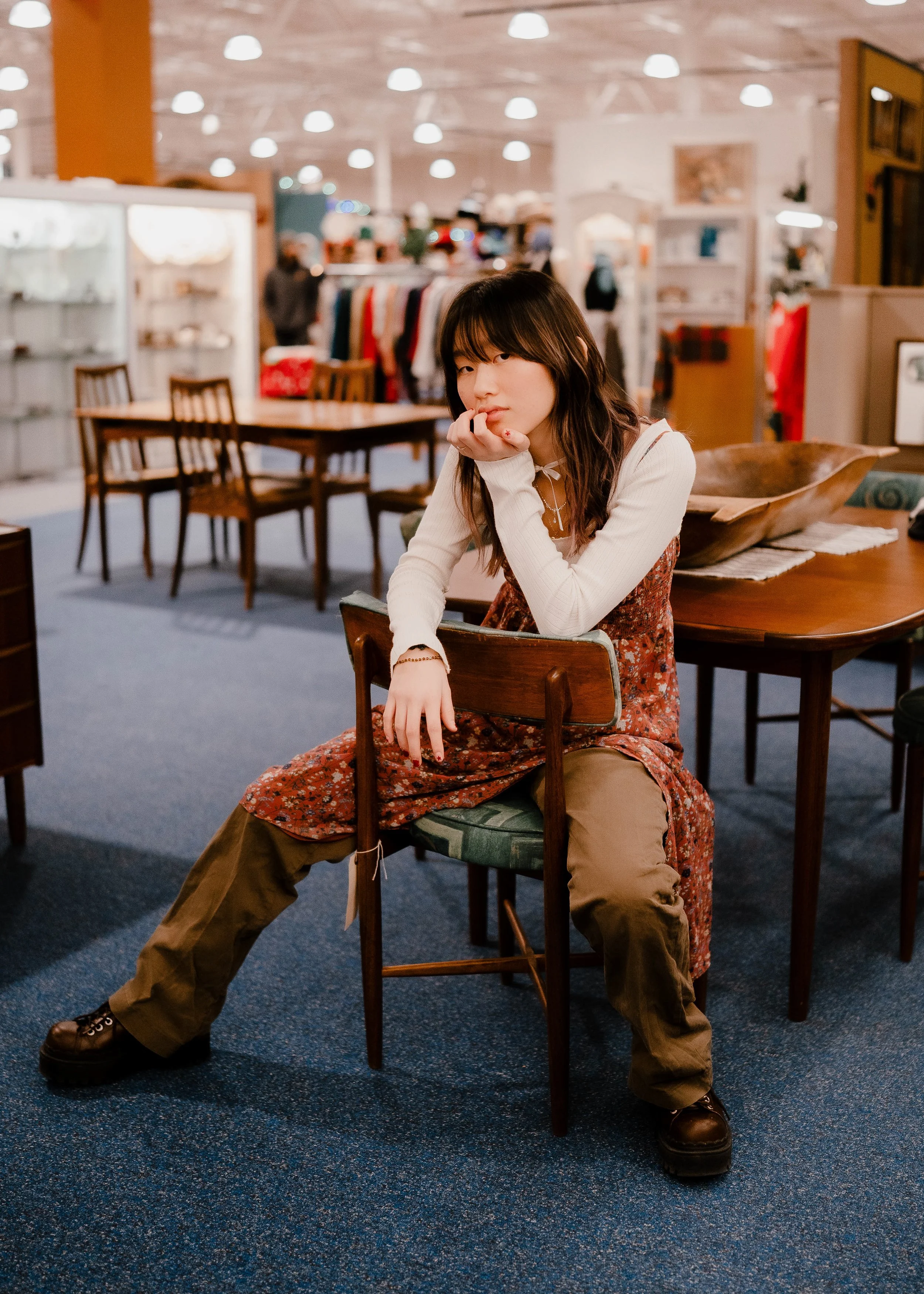 A young woman with long dark hair sitting on a wooden chair in a store or shopping mall. She is wearing a white long-sleeve shirt, a floral dress, and brown pants with black shoes. She is resting her chin on her hand, looking at the camera with a tho
