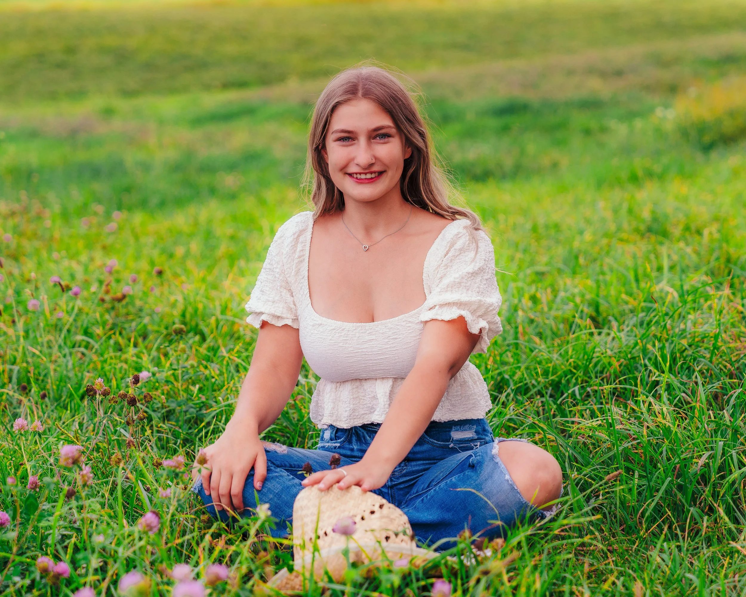 A young woman with long blonde hair smiling, sitting cross-legged in a grassy field with purple flowers, wearing a white ruffled blouse and ripped jeans, holding a straw hat.