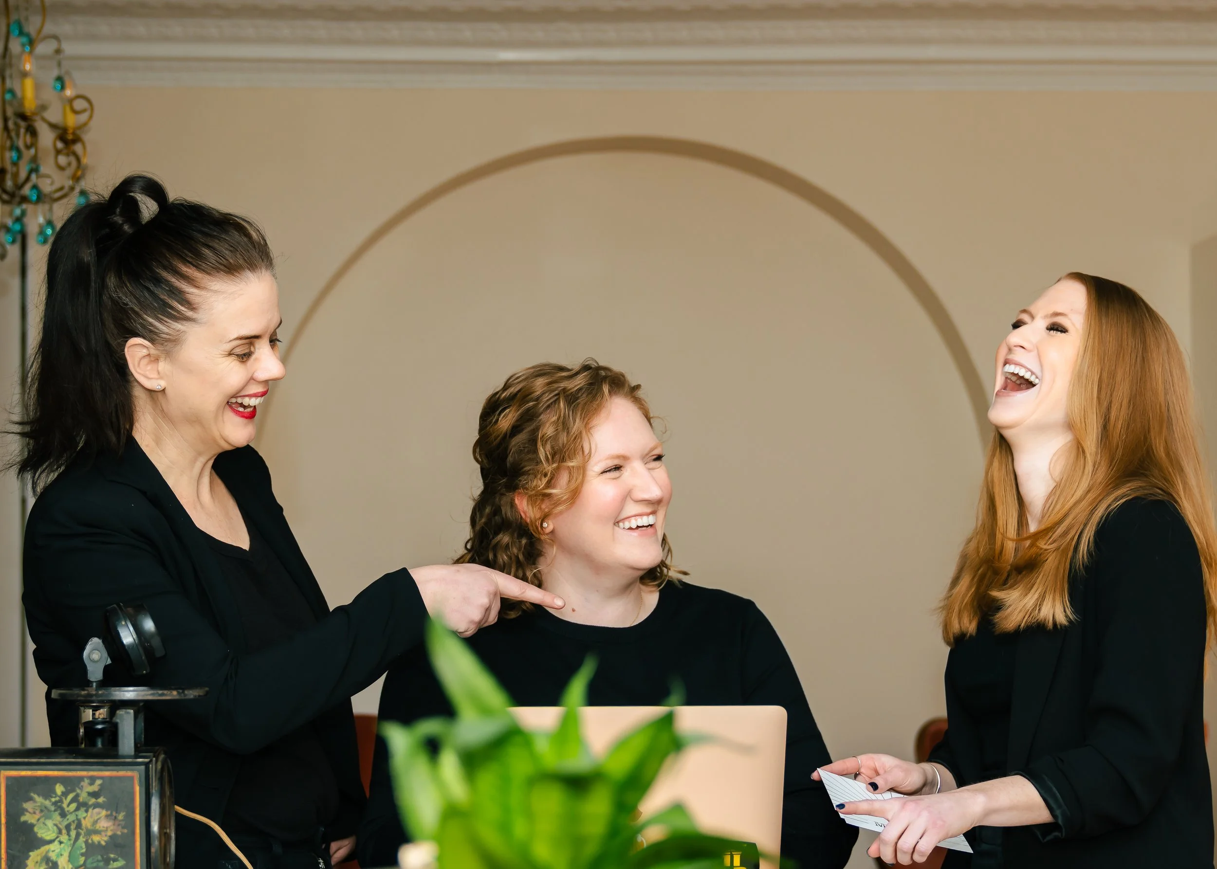 Three women at a table, laughing and talking, with a laptop and a potted plant in the foreground.
