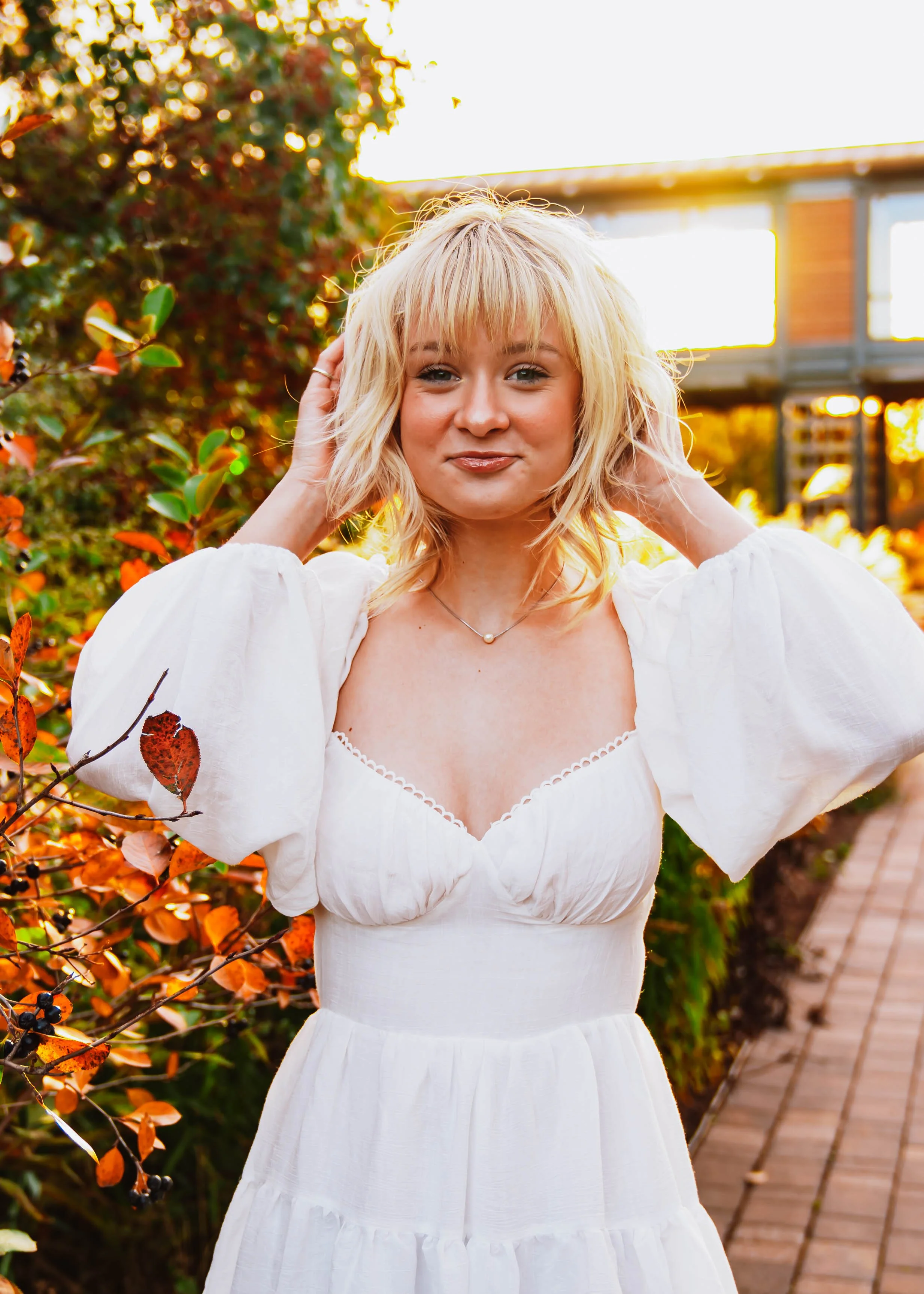 A woman with blonde hair wearing a white dress standing outdoors among autumn foliage, with a wooden walkway and a building in the background.