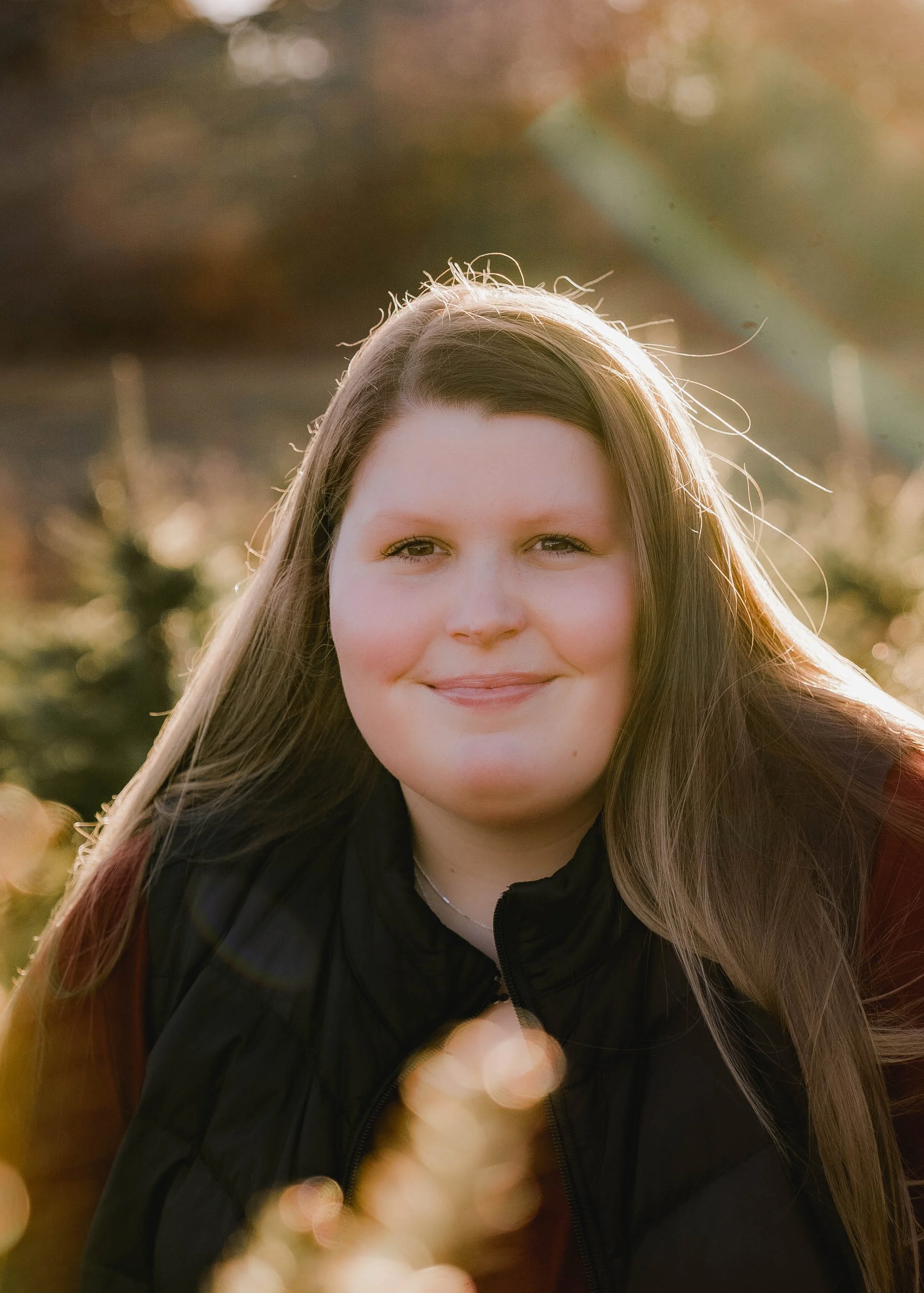 A young woman with long brown hair smiling outdoors during golden hour with sunlight shining behind her and bokeh of trees in the background.