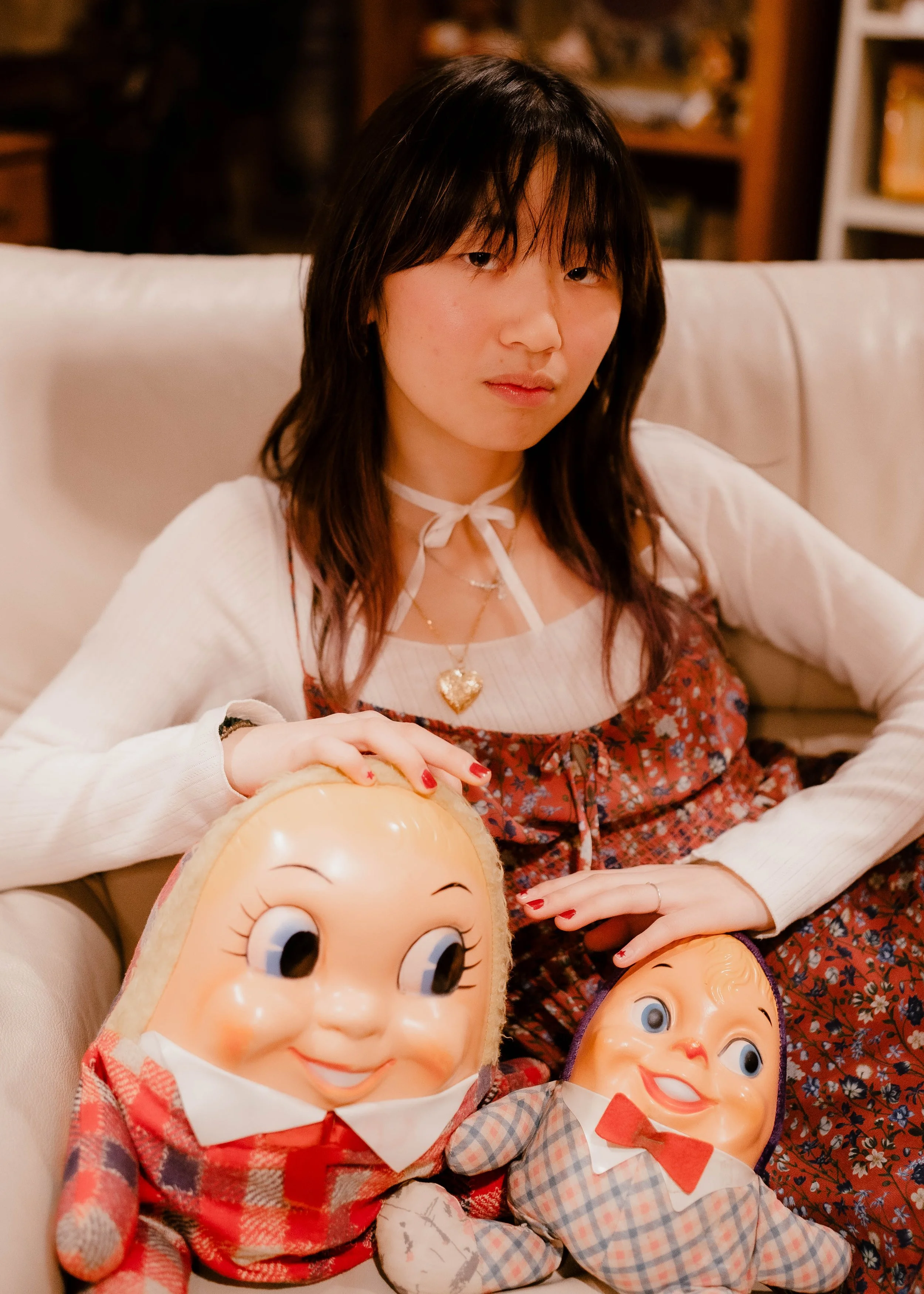 A young woman sitting on a white couch, holding two vintage plush dolls with large heads and smiling faces. She has dark, shoulder-length hair and is wearing a white sweater under a patterned dress, accessorized with necklaces and a ribbon around her