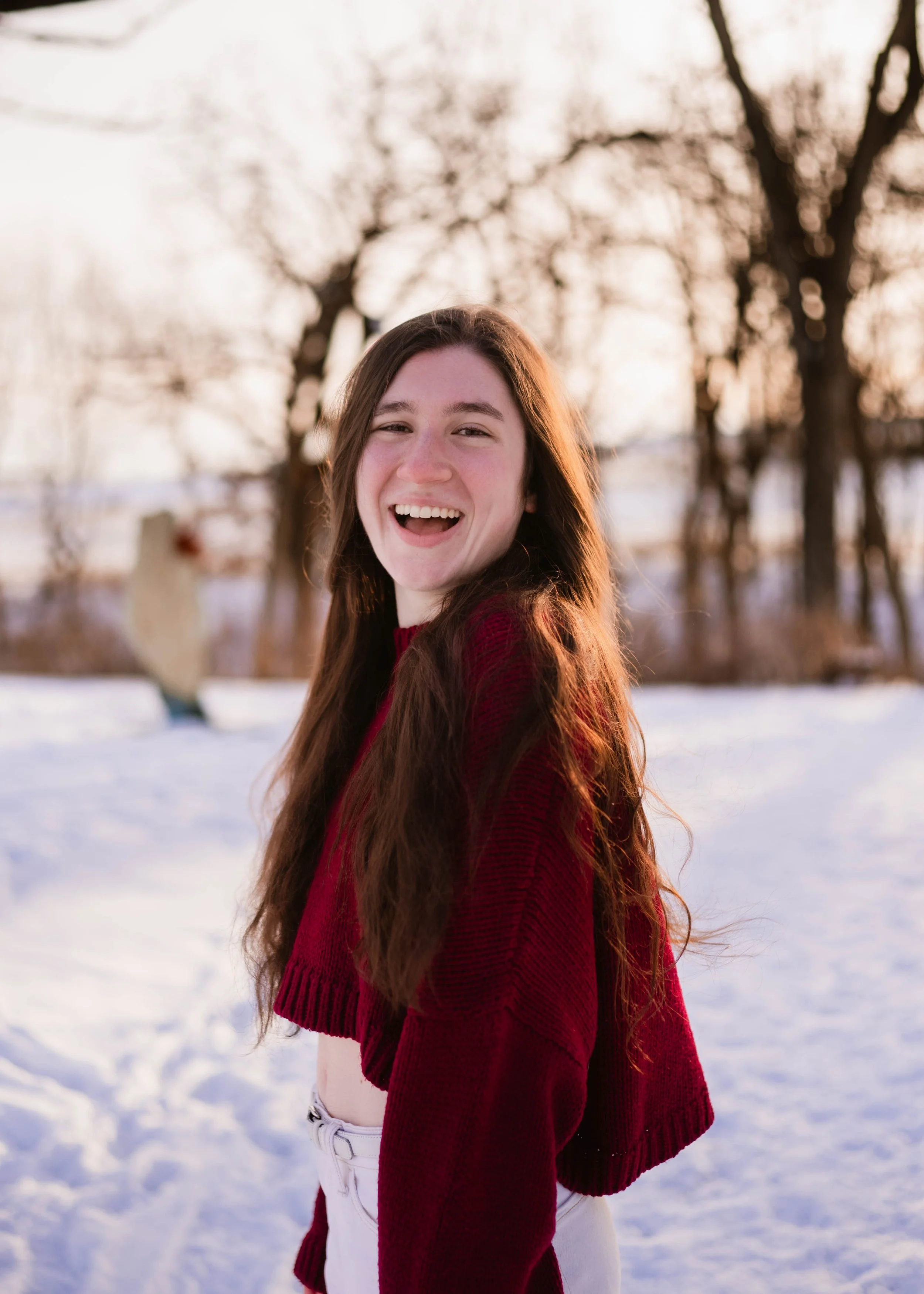 A young woman with long brown hair, smiling and laughing outdoors in a snowy landscape during sunset, wearing a red sweater and white pants.