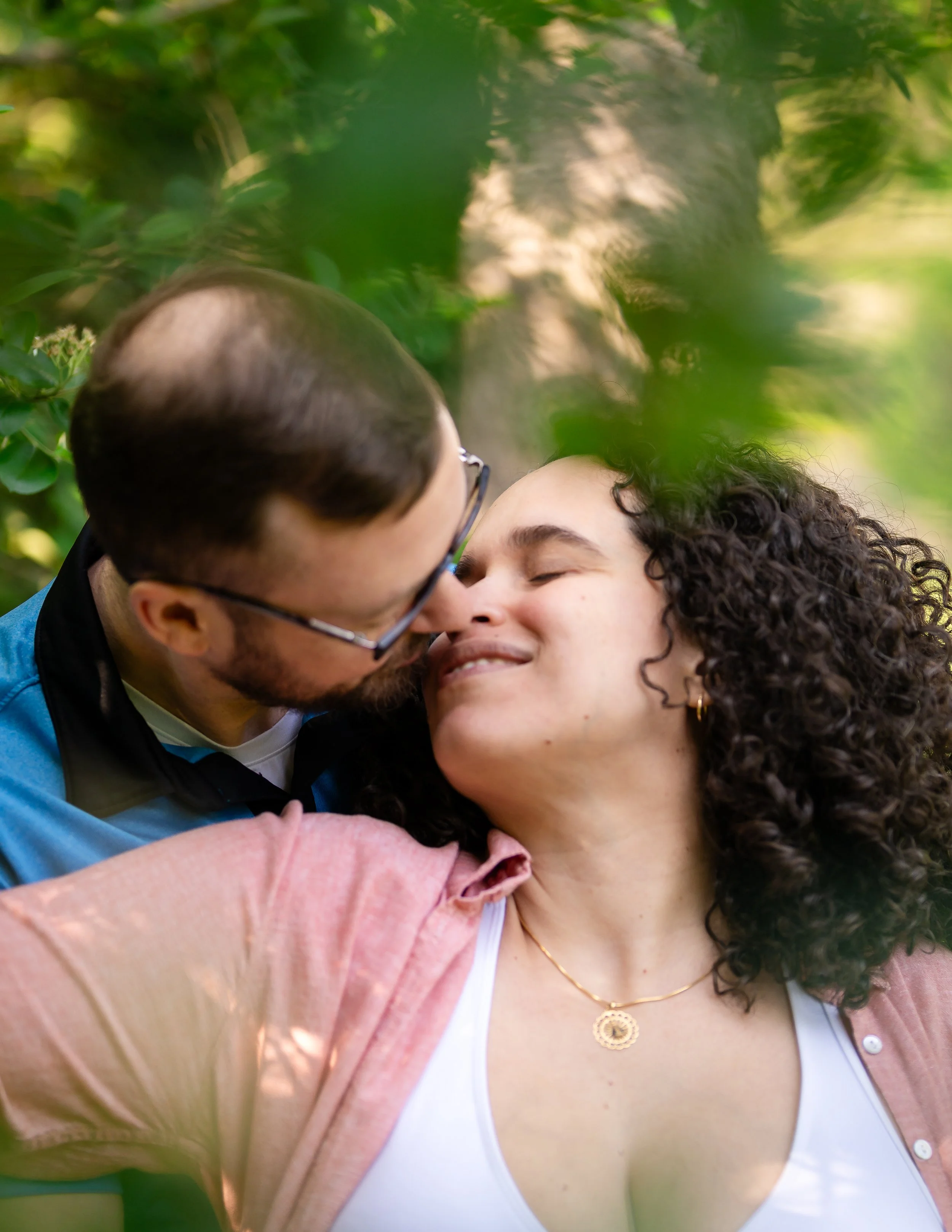 A couple sharing a tender moment outdoors, with the man leaning in to kiss the woman on the nose, surrounded by green foliage.