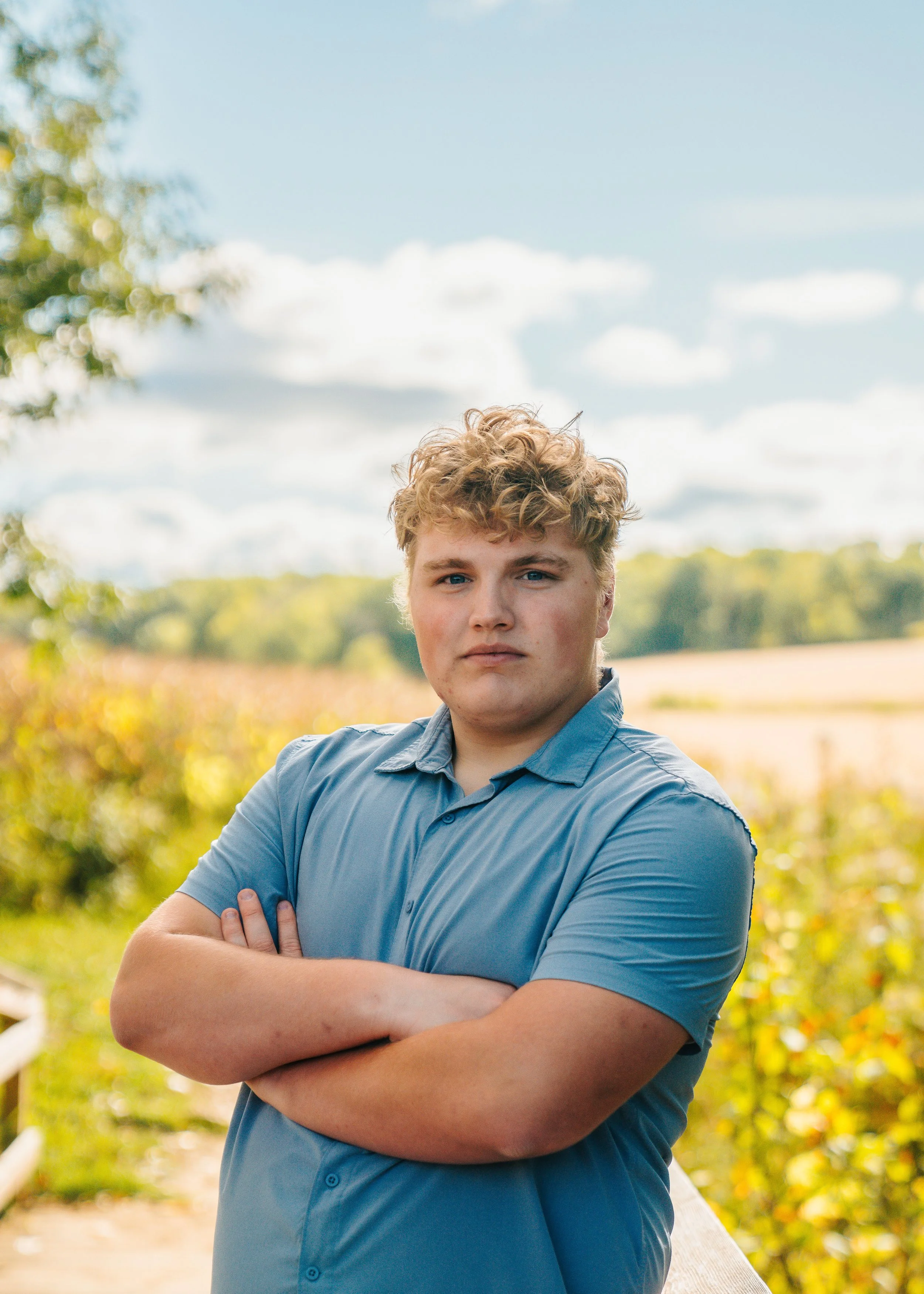 A young man with curly blond hair, wearing a light blue shirt, stands outdoors with his arms crossed, on a bridge or walkway in a rural setting with farmland and trees in the background.