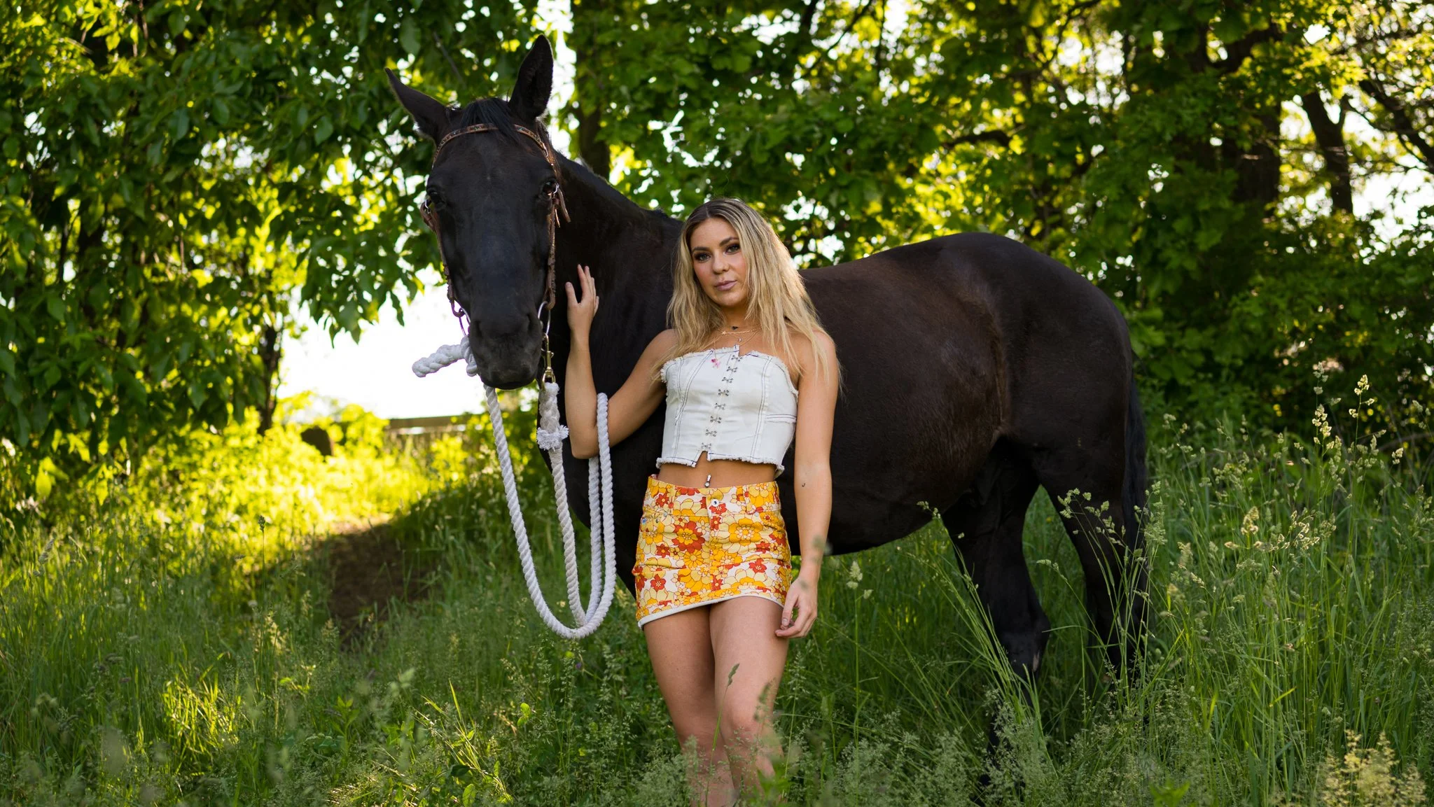 A young woman stands next to a black horse in a grassy, green outdoor setting, holding the horse's halter.