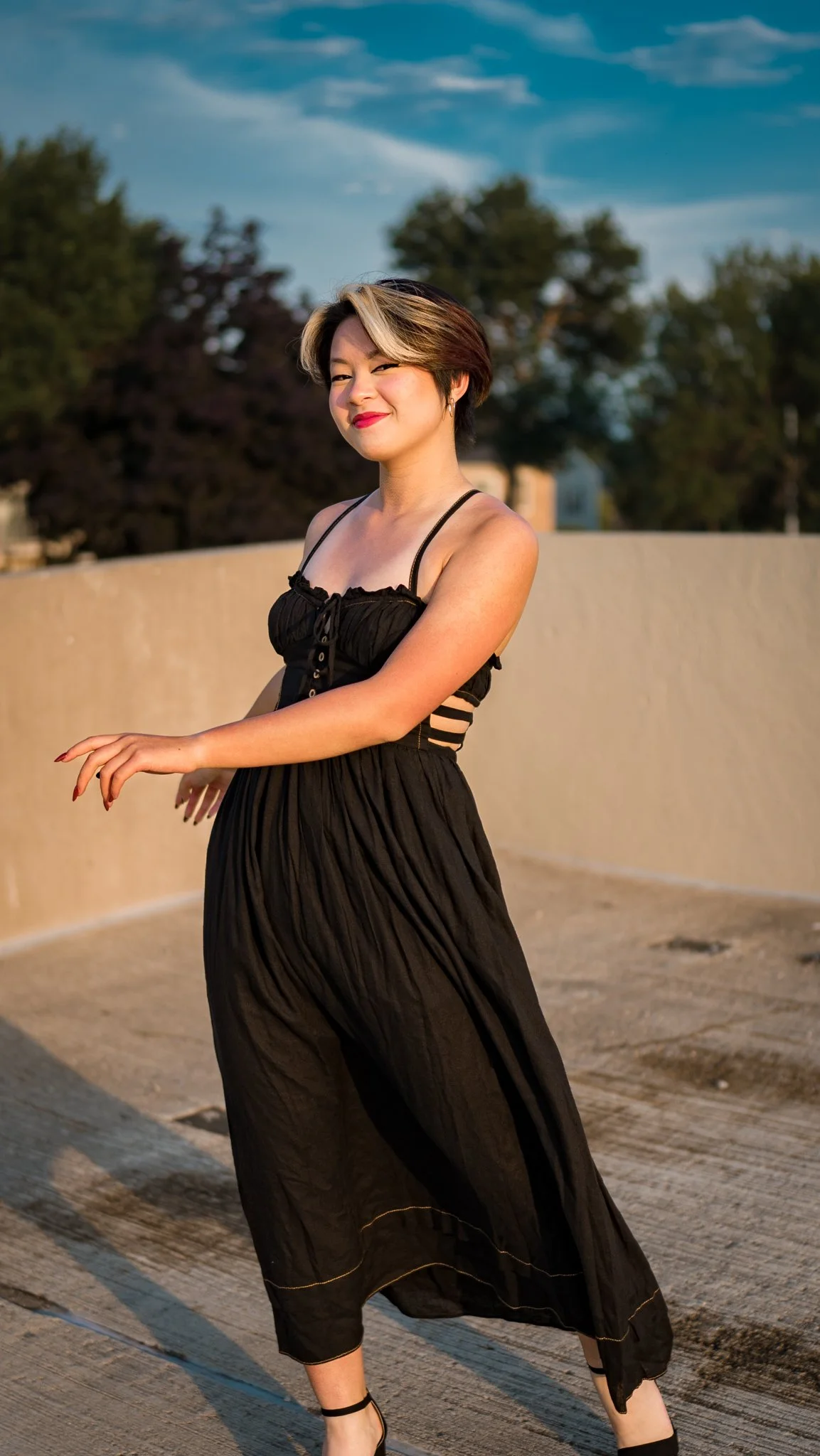 A woman in a black dress standing outdoors, smiling and squinting in the sunlight with trees and a blue sky in the background.