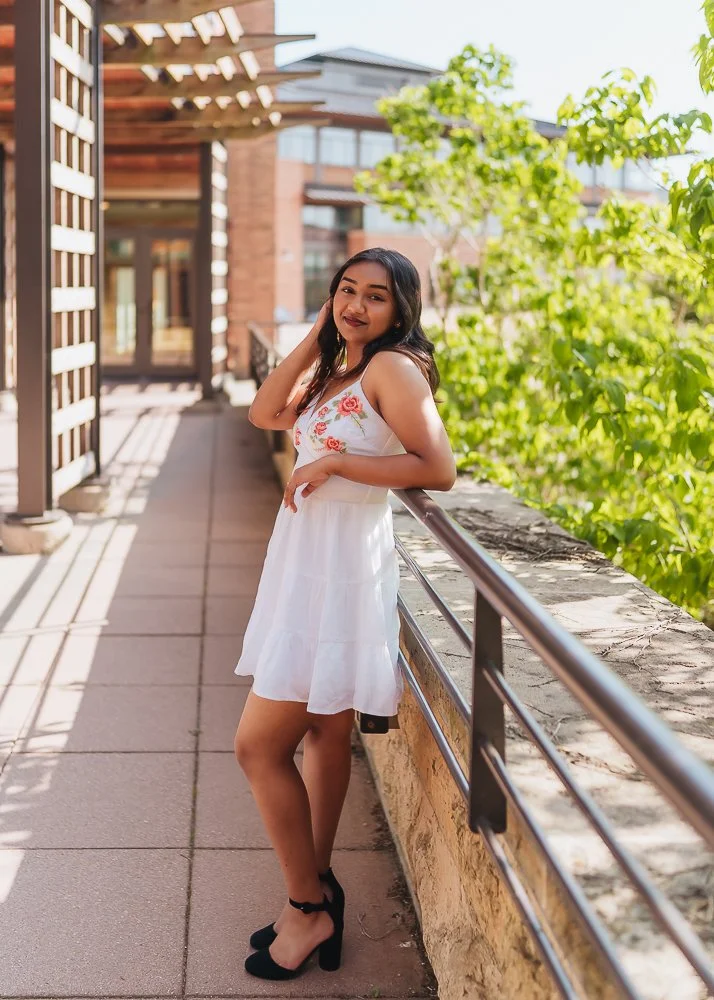 A young woman in a white dress with floral embroidery standing on a balcony with greenery in the background.