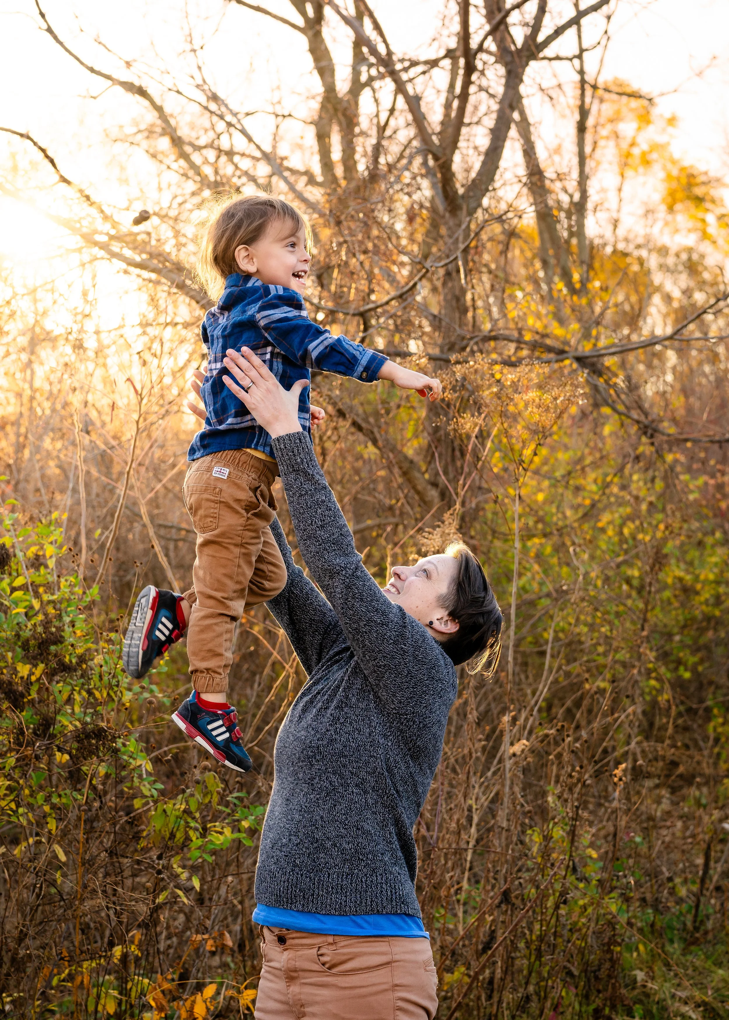 A woman lifting a young boy into the air outdoors during autumn, with trees and fall foliage visible in the background.