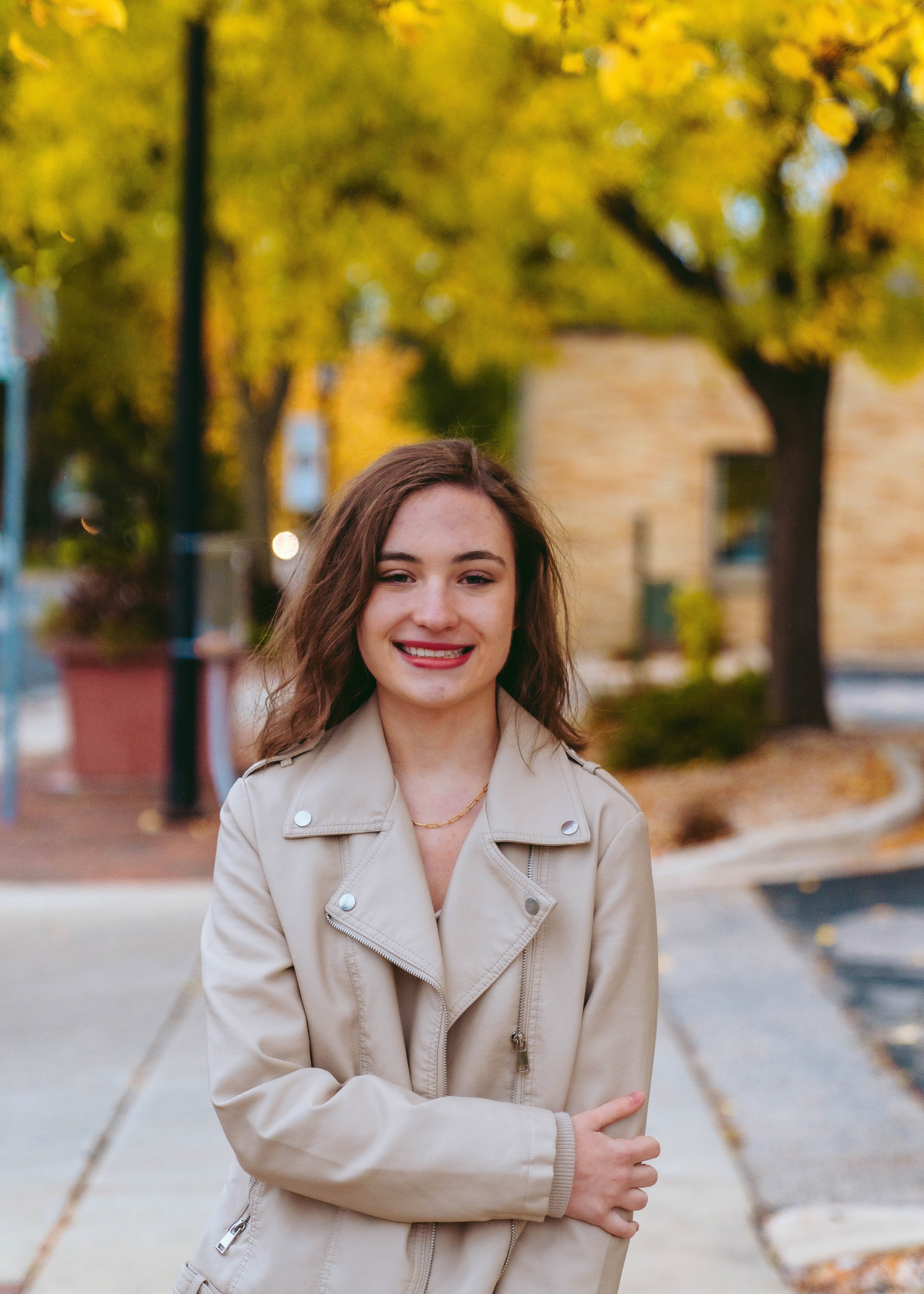 Young woman with brown hair wearing a beige leather jacket, smiling outdoors with trees and buildings in autumn background.