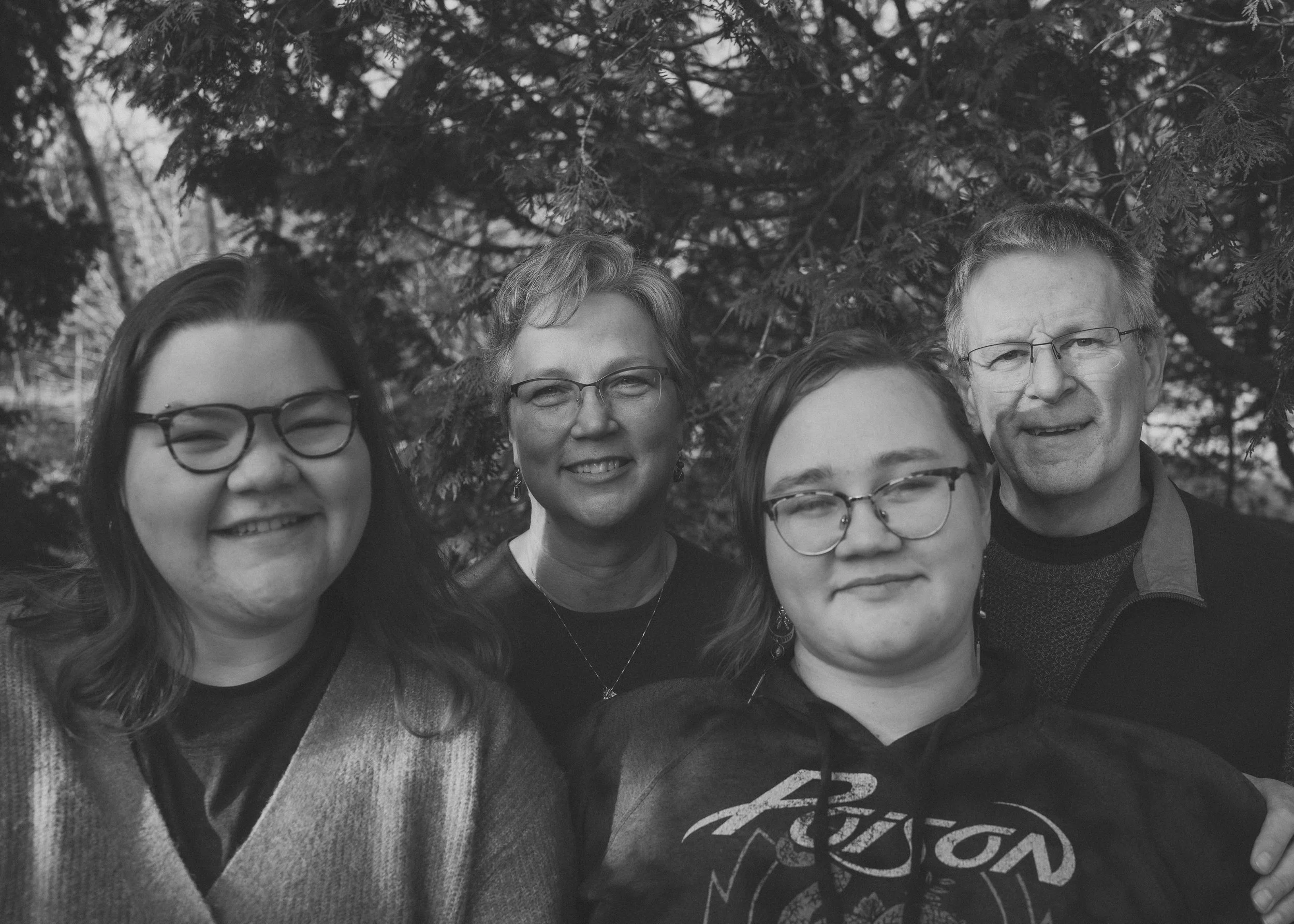 Black and white photo of four people outdoors, standing in front of trees. The group includes two women and two men, all wearing glasses and smiling at the camera.