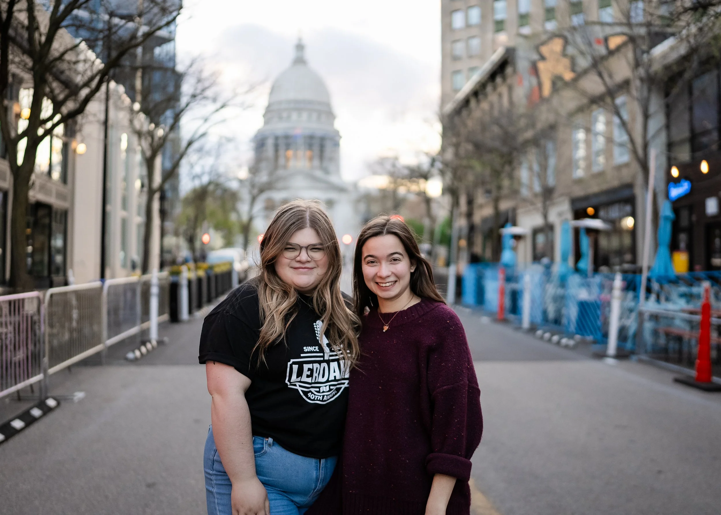 Two young women standing on a city street with the US Capitol building in the background, smiling at the camera.
