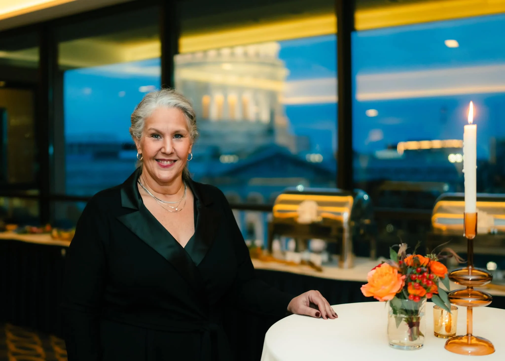 An older woman with grey hair smiling at a dinner event, standing next to a table with a lit candle and a floral arrangement, in a room with large windows showing a cityscape at dusk or nighttime.