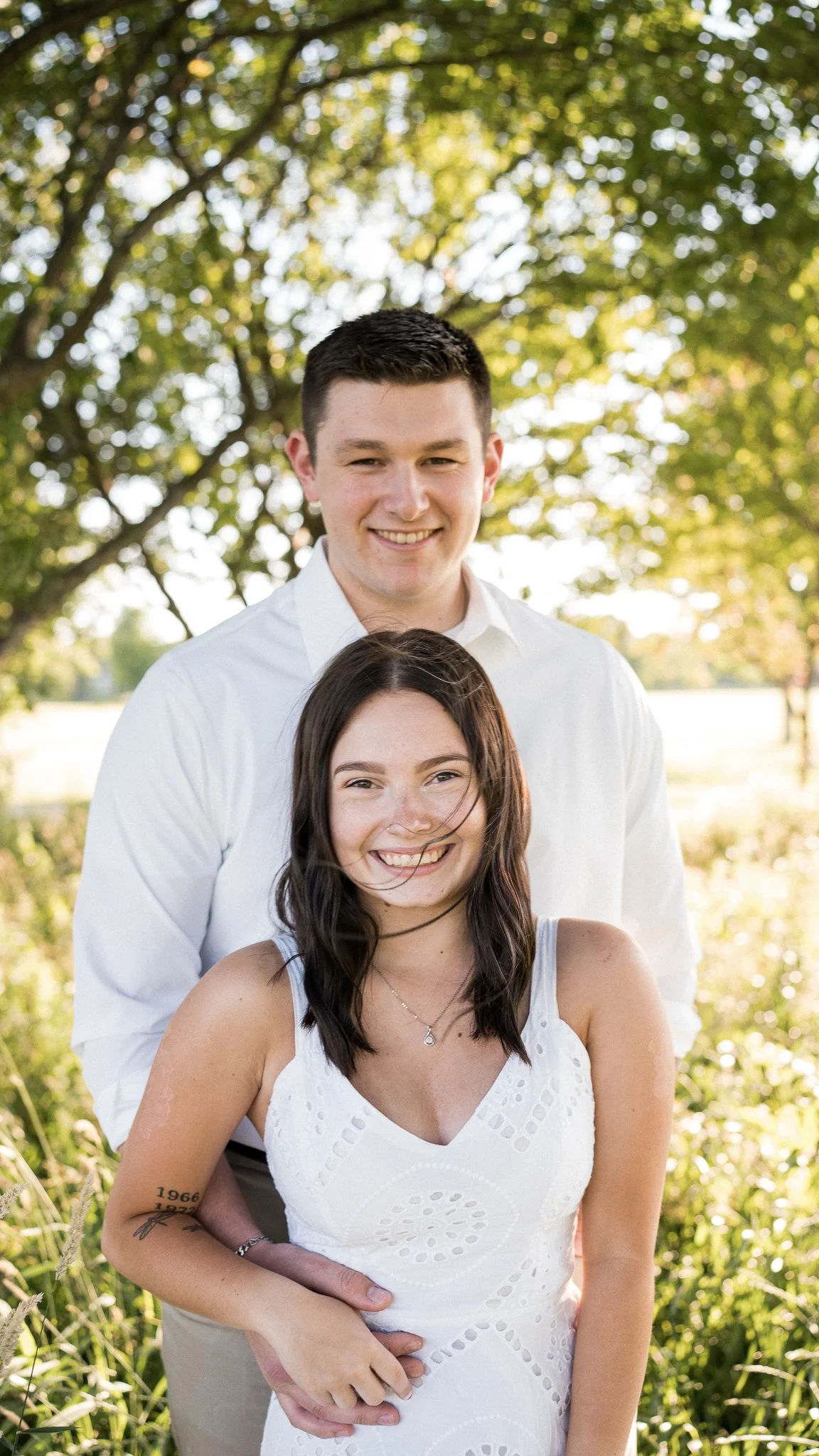 A young couple smiling outdoors in a park, standing close with the man behind the woman, both dressed in white.