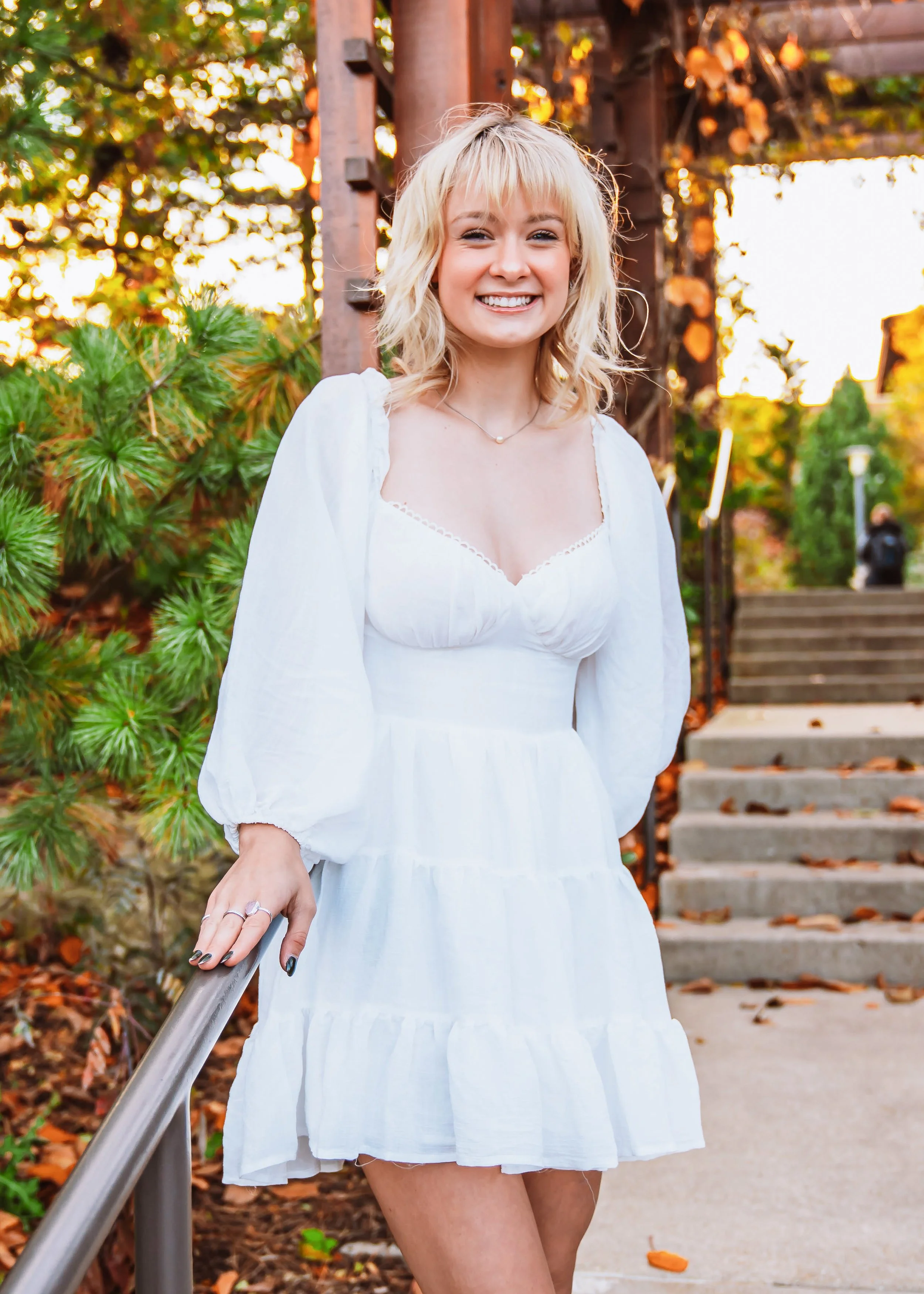 A young woman with blonde, wavy hair smiling and wearing a white dress outdoors during fall, with stairs and trees with orange leaves in the background.
