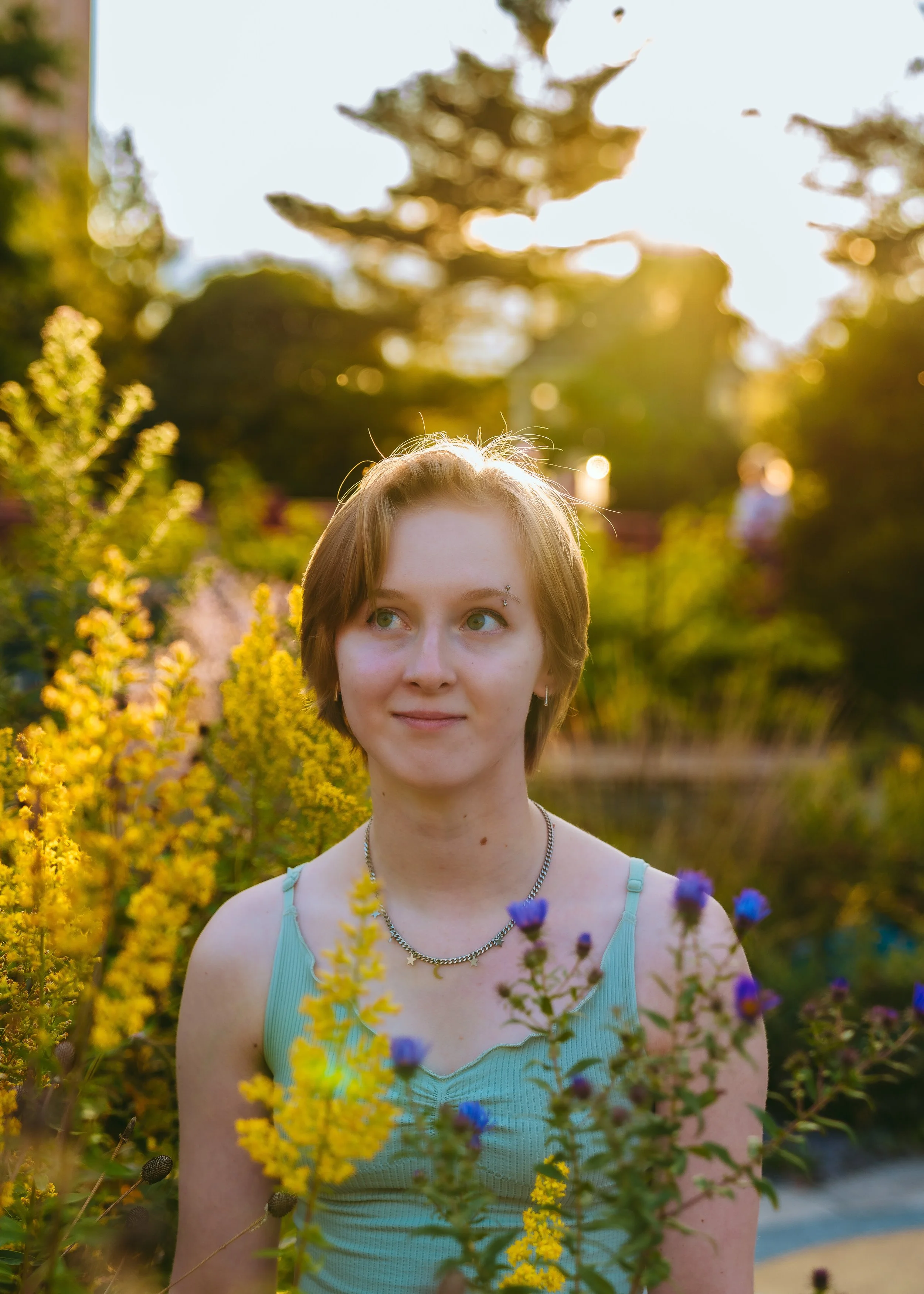 A young woman with short brown hair and a linked necklace is standing among colorful flowers in a garden during golden hour, with sunlight shining behind her.