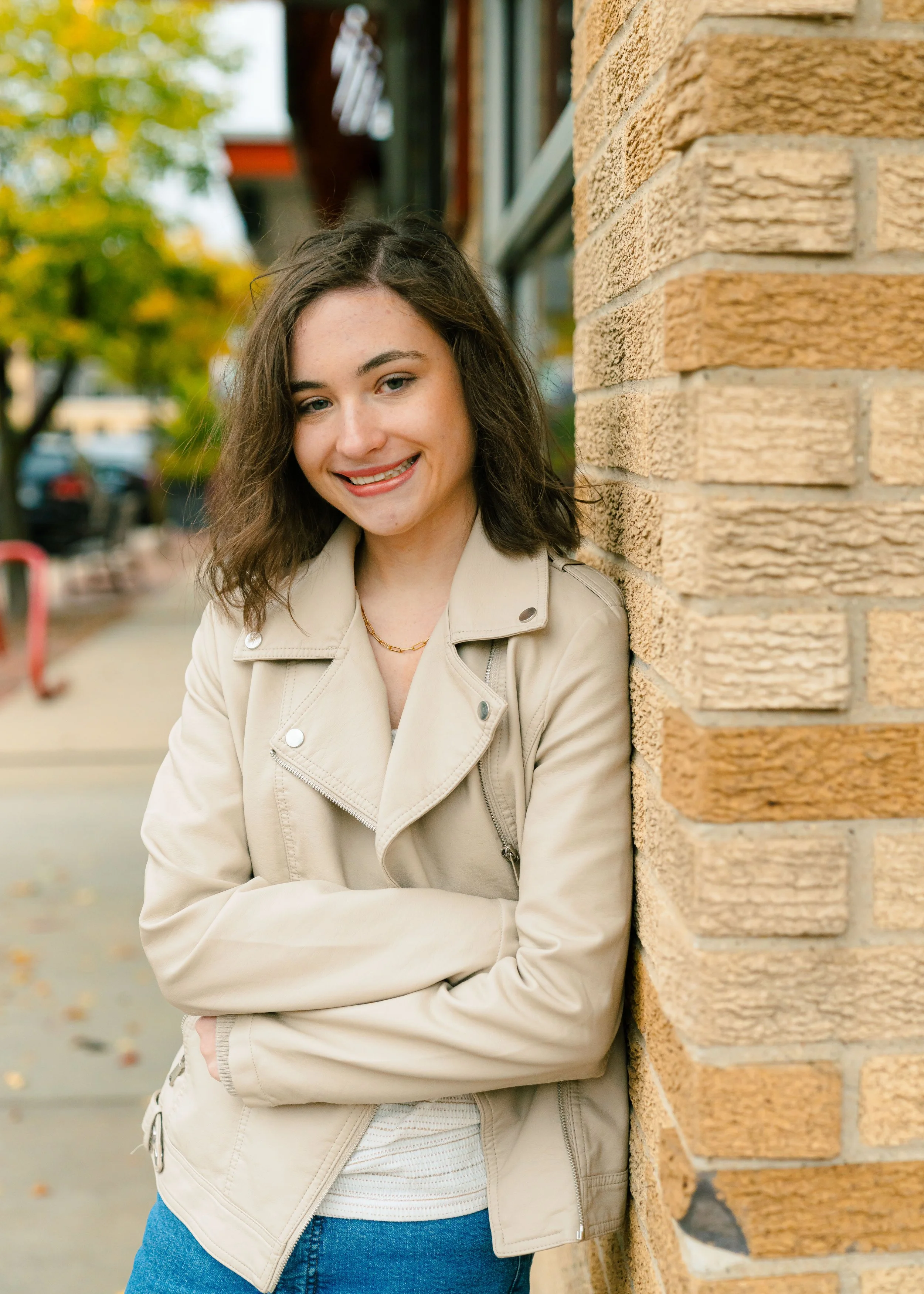 A young woman with shoulder-length brown hair, smiling, wearing a beige jacket, standing outdoors next to a brick wall with trees and a sidewalk in the background.