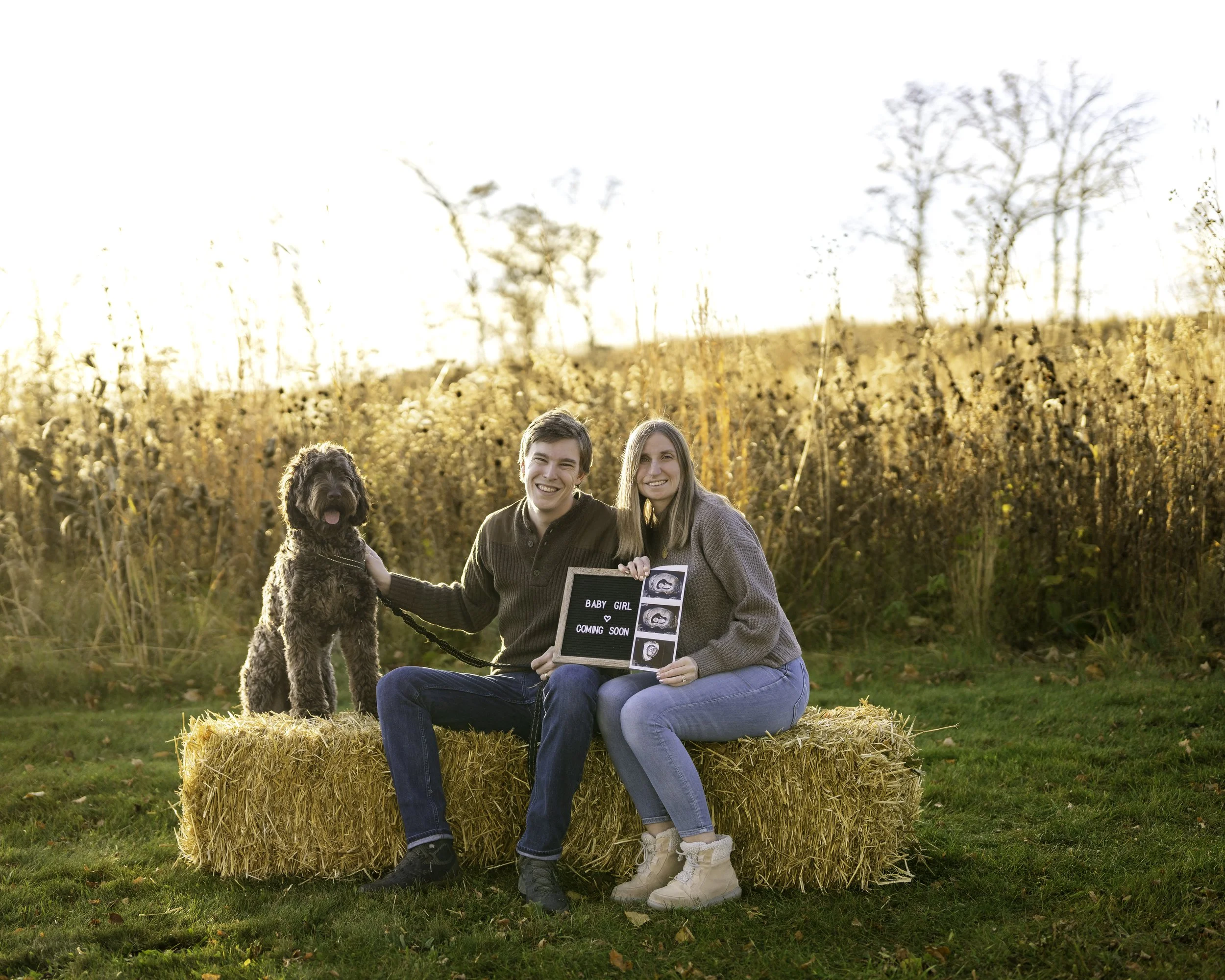 A happy couple sits on hay bales outdoors during autumn with their dog, holding a sign and ultrasound images announcing expecting a baby girl soon.