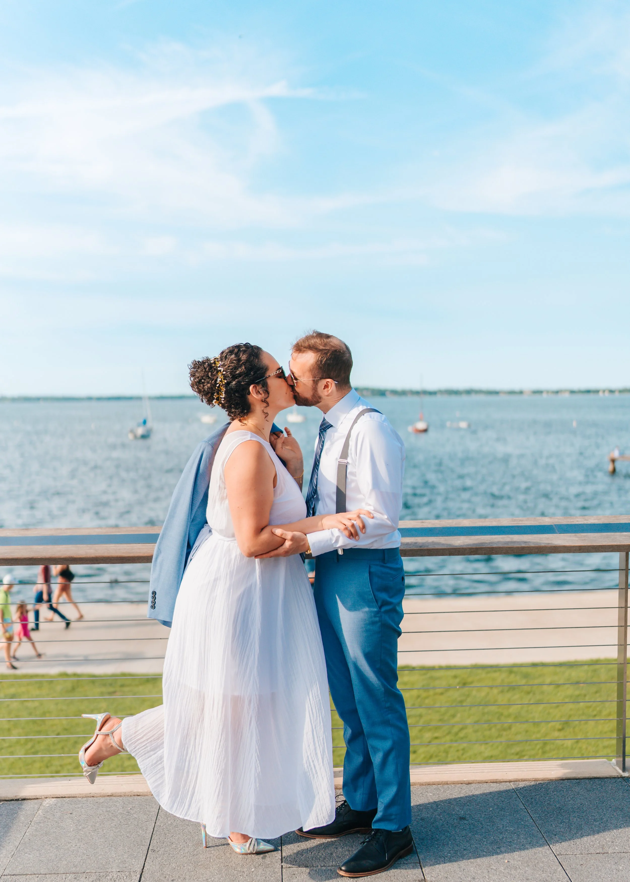 A couple sharing a kiss by the waterfront on a sunny day, with sailboats in the background.