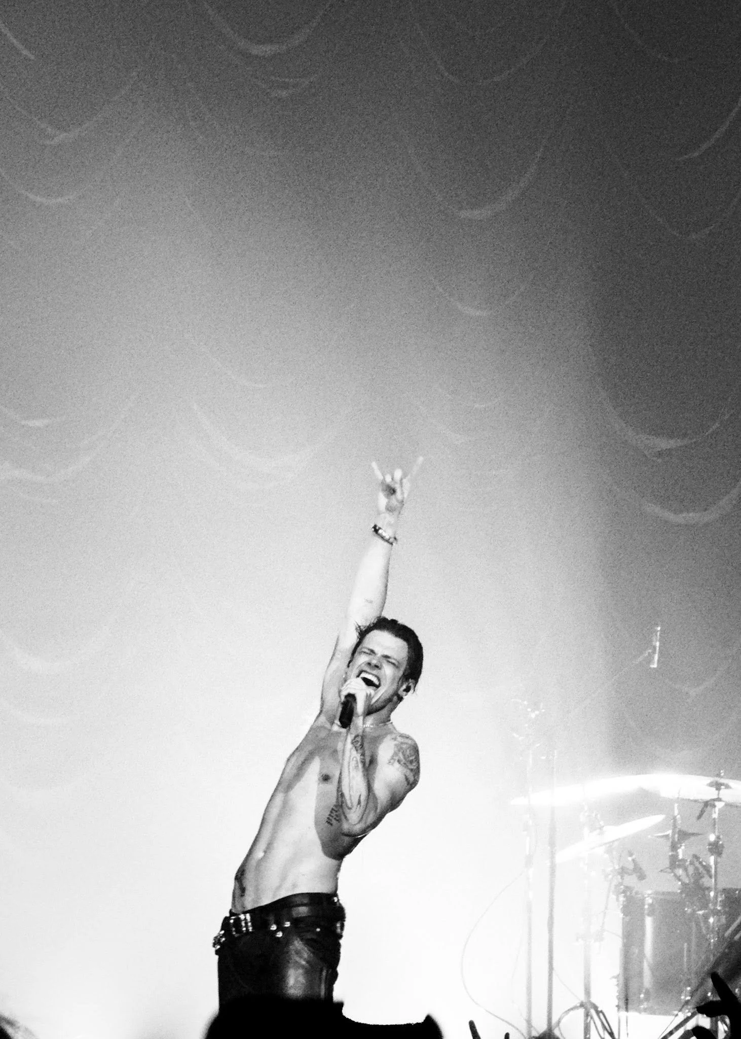 A shirtless man with tattoos singing into a microphone on stage, with his right arm raised and making a rock gesture, and a drum set visible in the background.