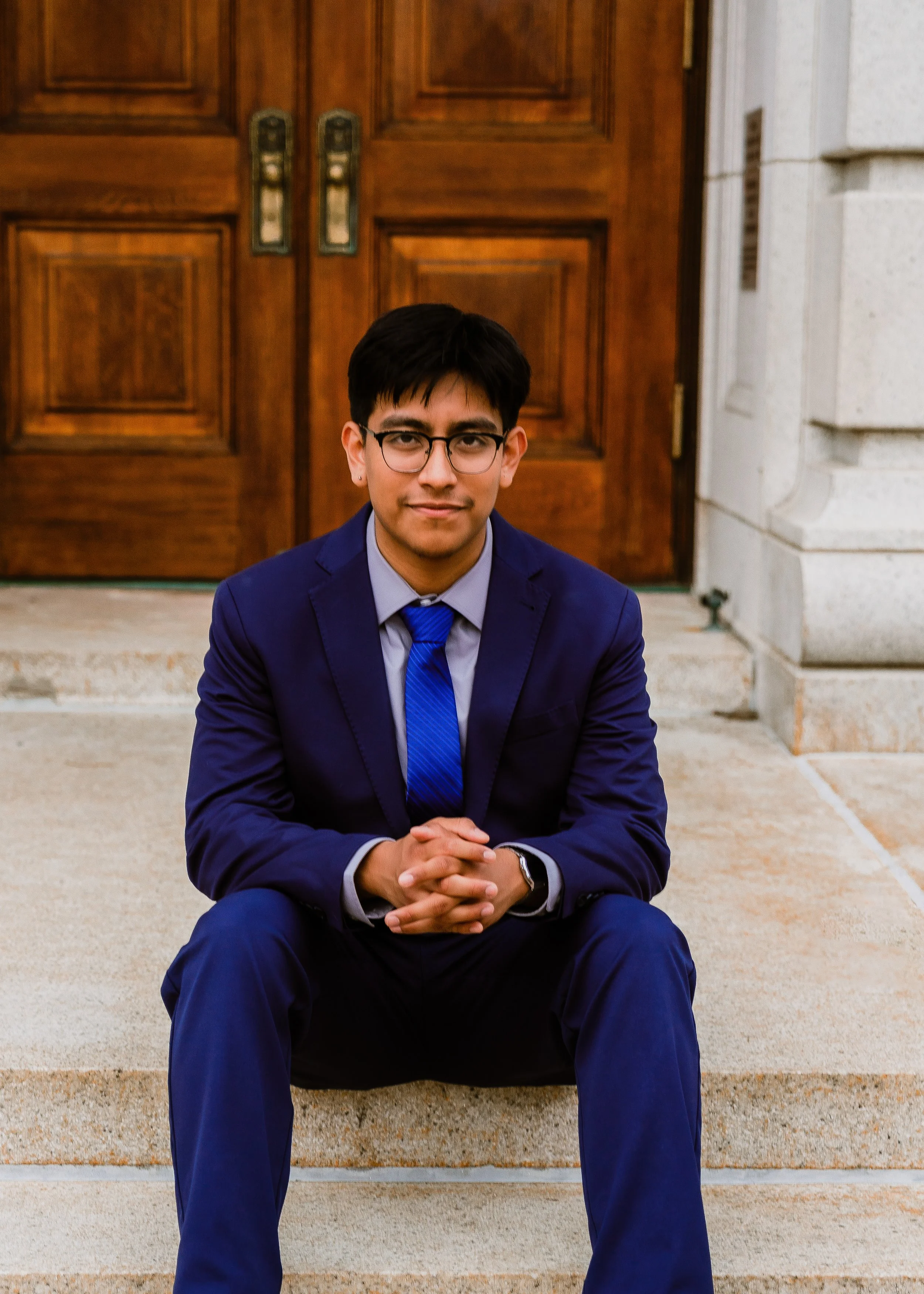 A young man wearing glasses, a dark blue suit, and a matching blue tie sitting on concrete steps in front of wooden double doors of a building.