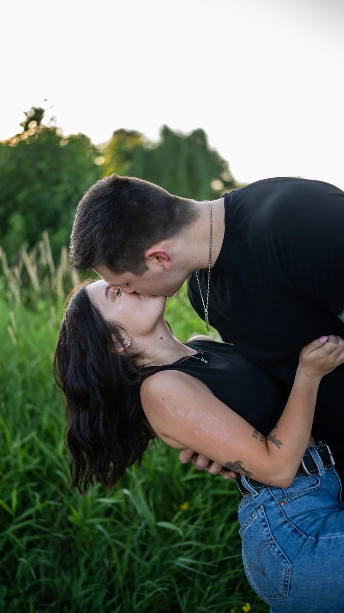 A young couple sharing a kiss outdoors in a grassy field with trees in the background.