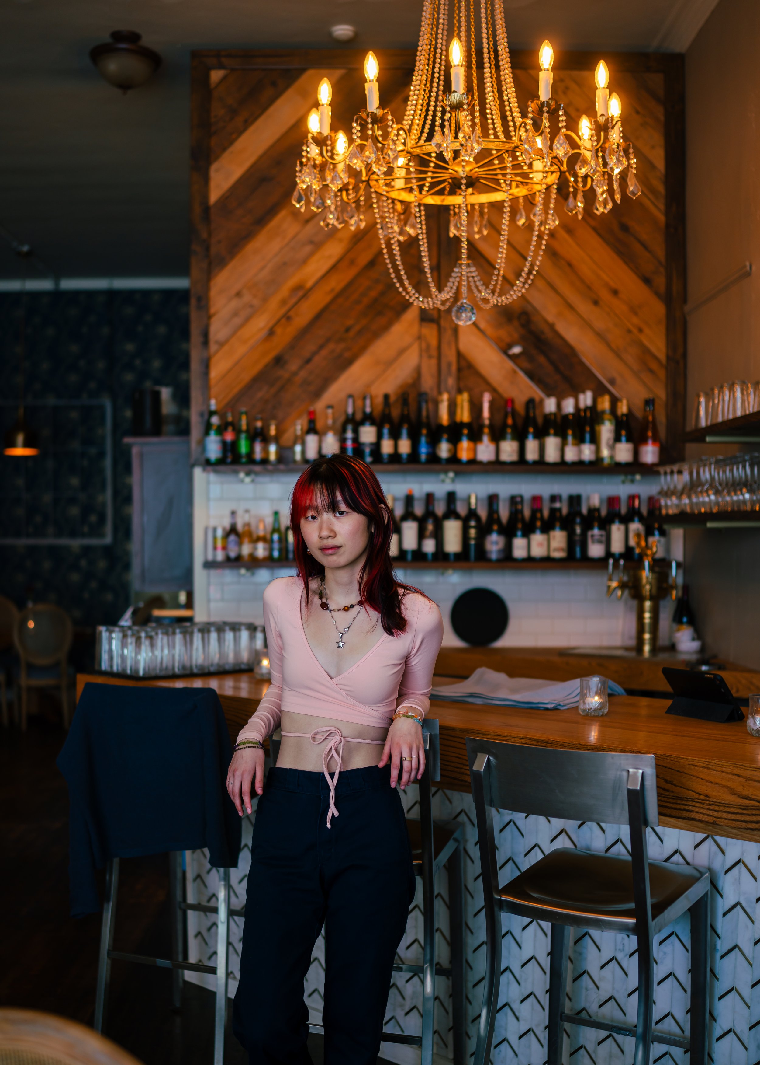 A woman with red hair in a pink crop top and black pants standing at a bar in a dimly lit restaurant with a chandelier and bottles of wine in the background.