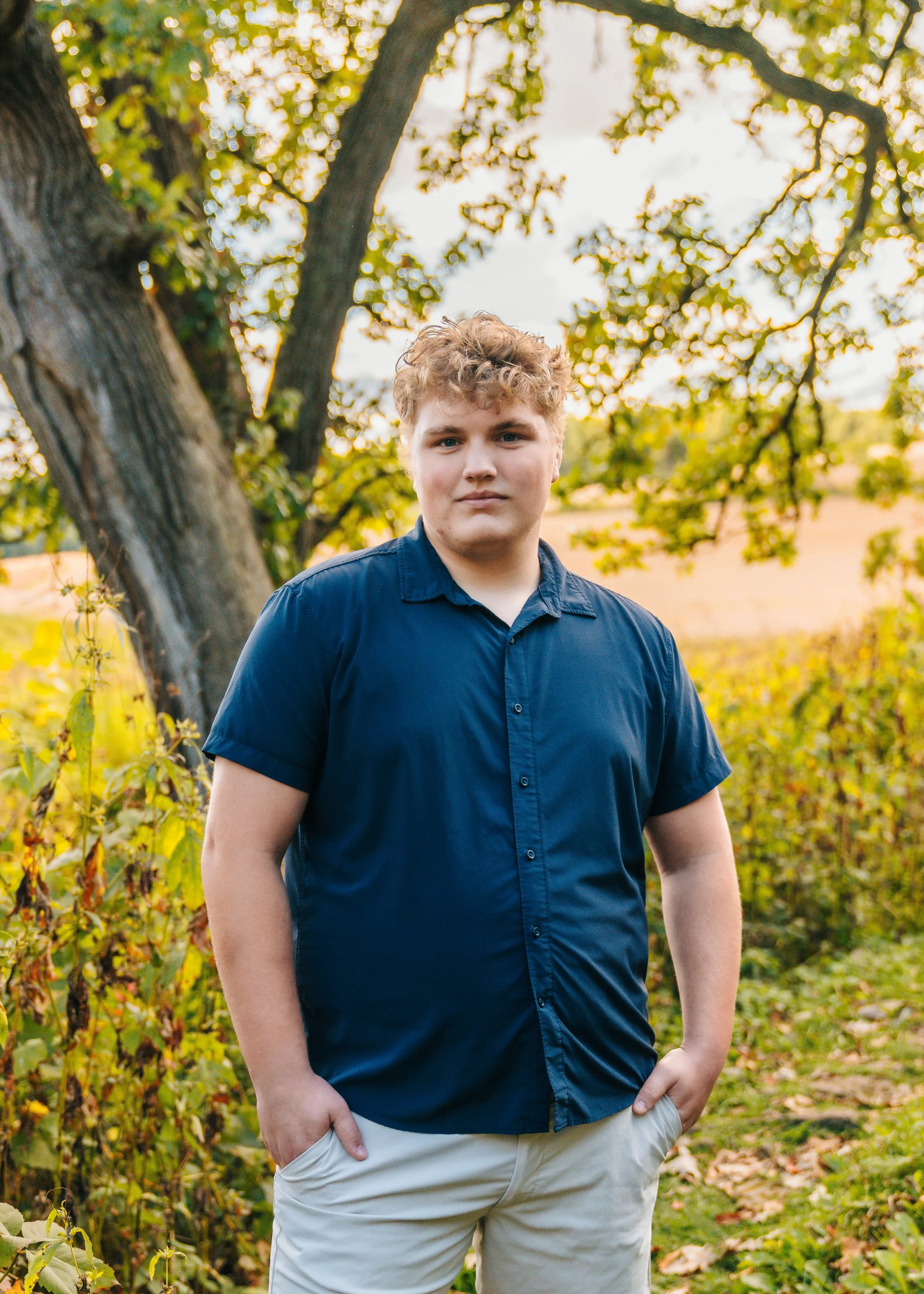 A young man with curly blonde hair wearing a navy blue shirt and beige pants standing outdoors in a natural setting with trees and foliage in the background.