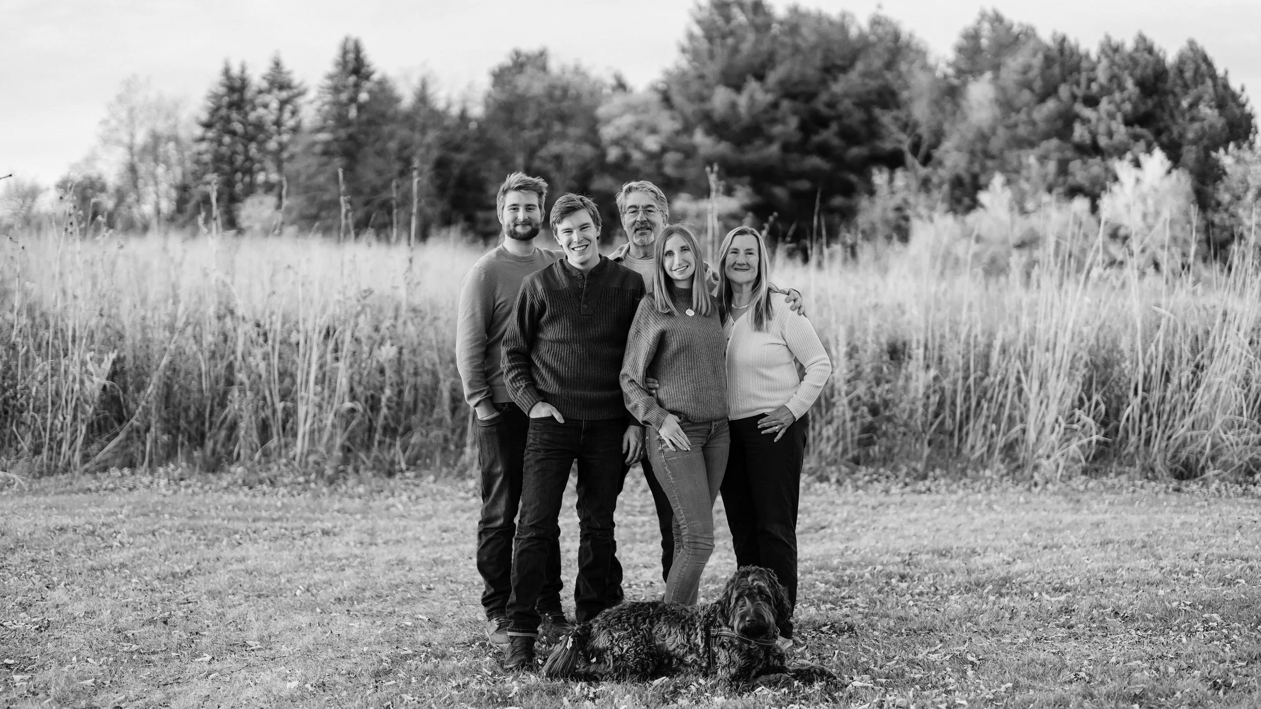 A black and white photo of six people standing outdoors in a field with tall grass and trees in the background, with a dog lying on the ground in front of them.