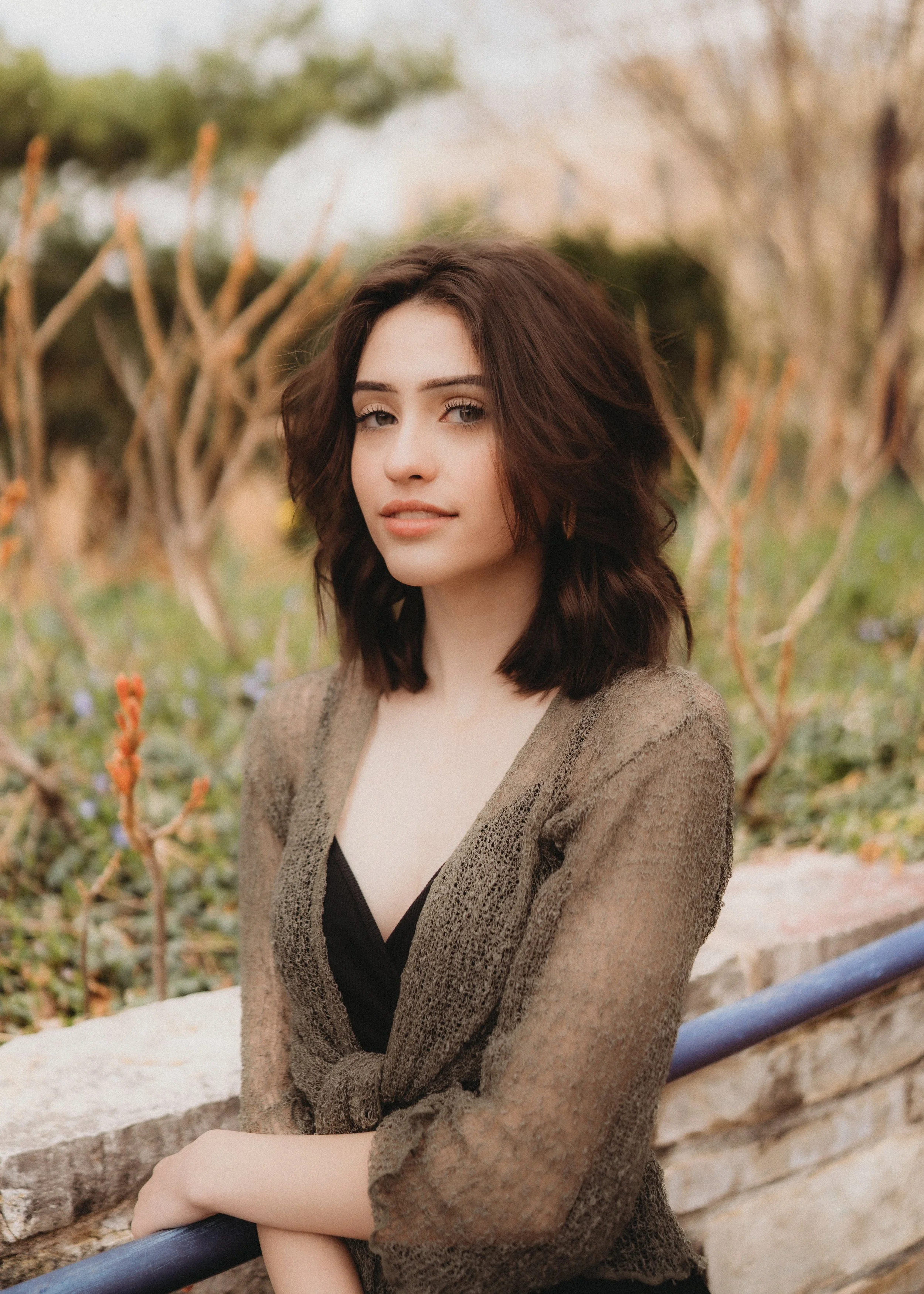 A young woman with shoulder-length brown hair and light skin posing outdoors near a stone wall and a shrub with thin branches, wearing a sheer brown blouse over a black top, looking at the camera.