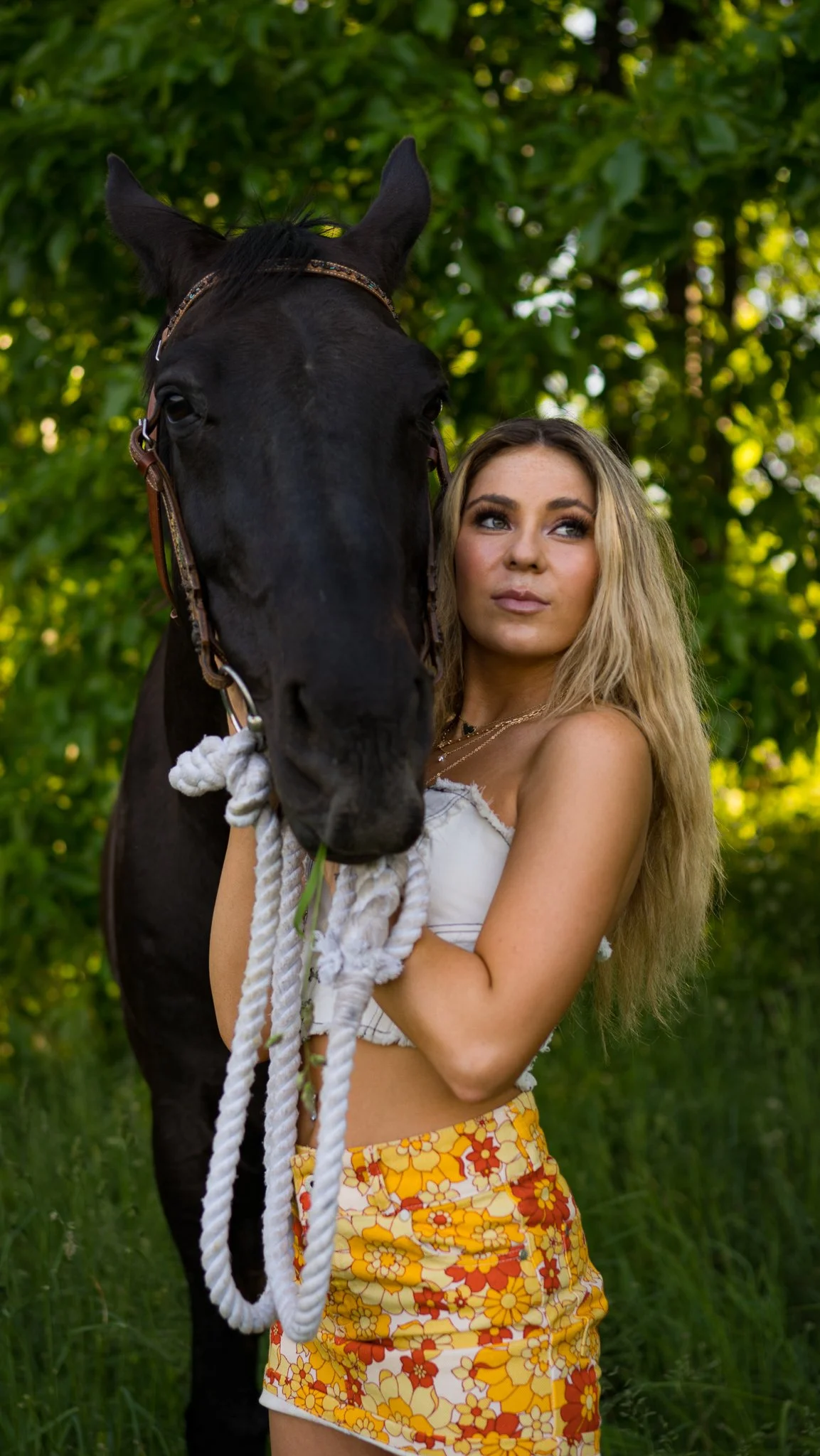 A woman with long blonde hair standing outdoors with a black horse, holding a rope attached to the horse's bridle, amidst green trees and grass.