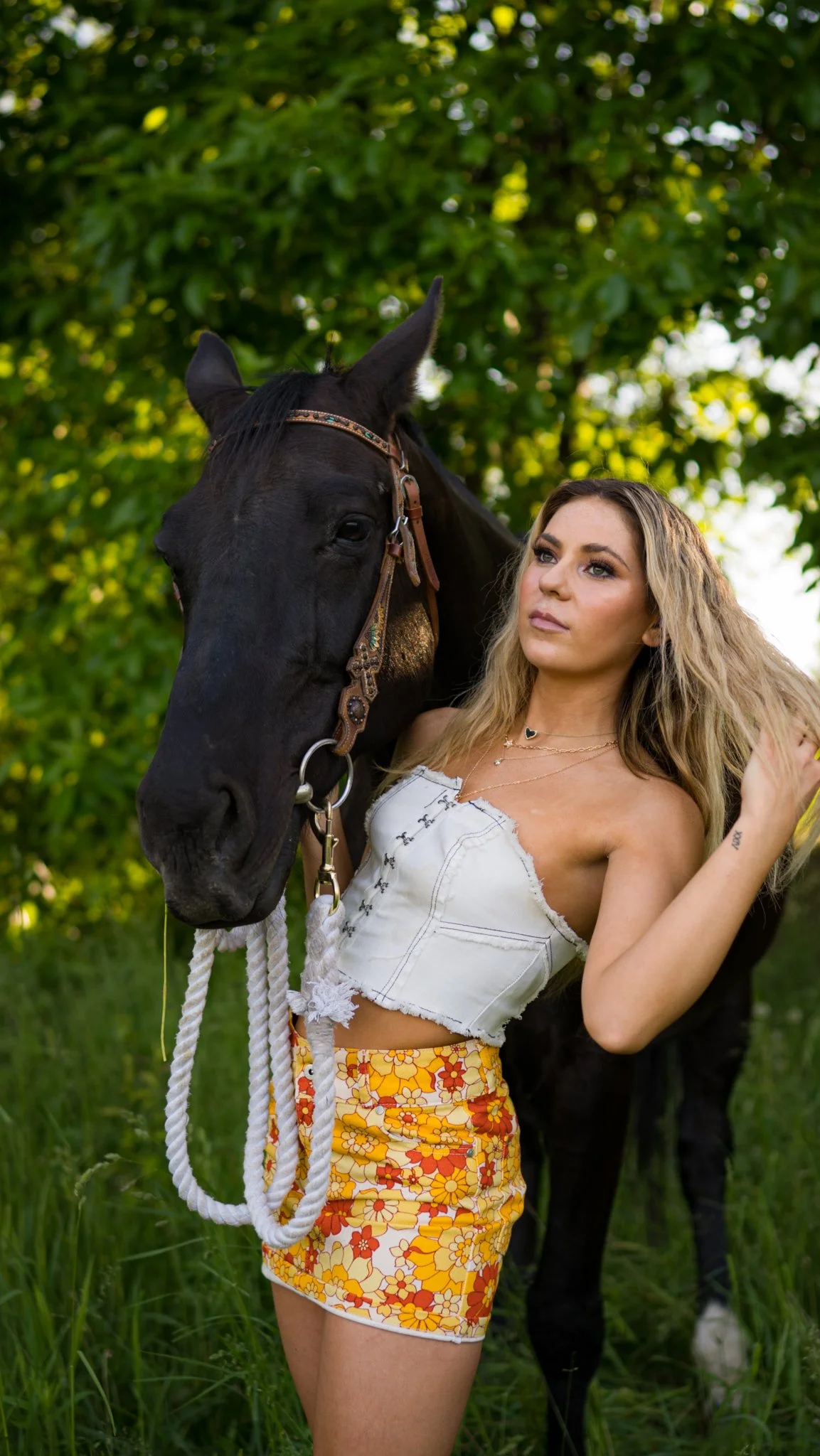 A woman with long blonde hair standing outdoors next to a black horse in a grassy area with green trees in the background.