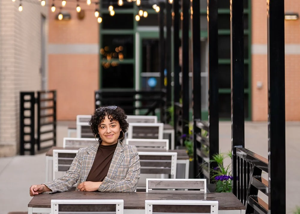 A woman with curly hair sitting at an outdoor table in a dining area with string lights, black railing, and a brick building in the background.