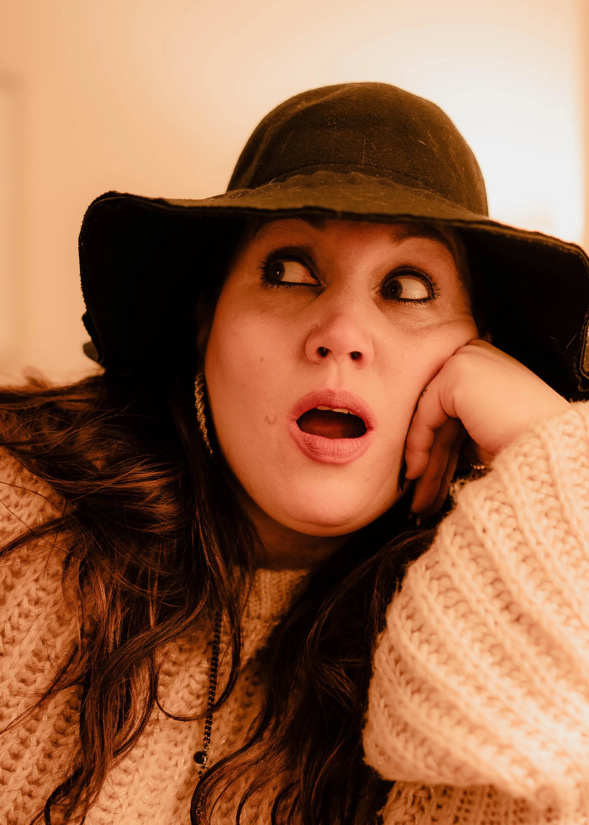 A woman wearing a large black hat and a beige sweater, resting her head on her hand with her mouth open and eyes looking to the side.