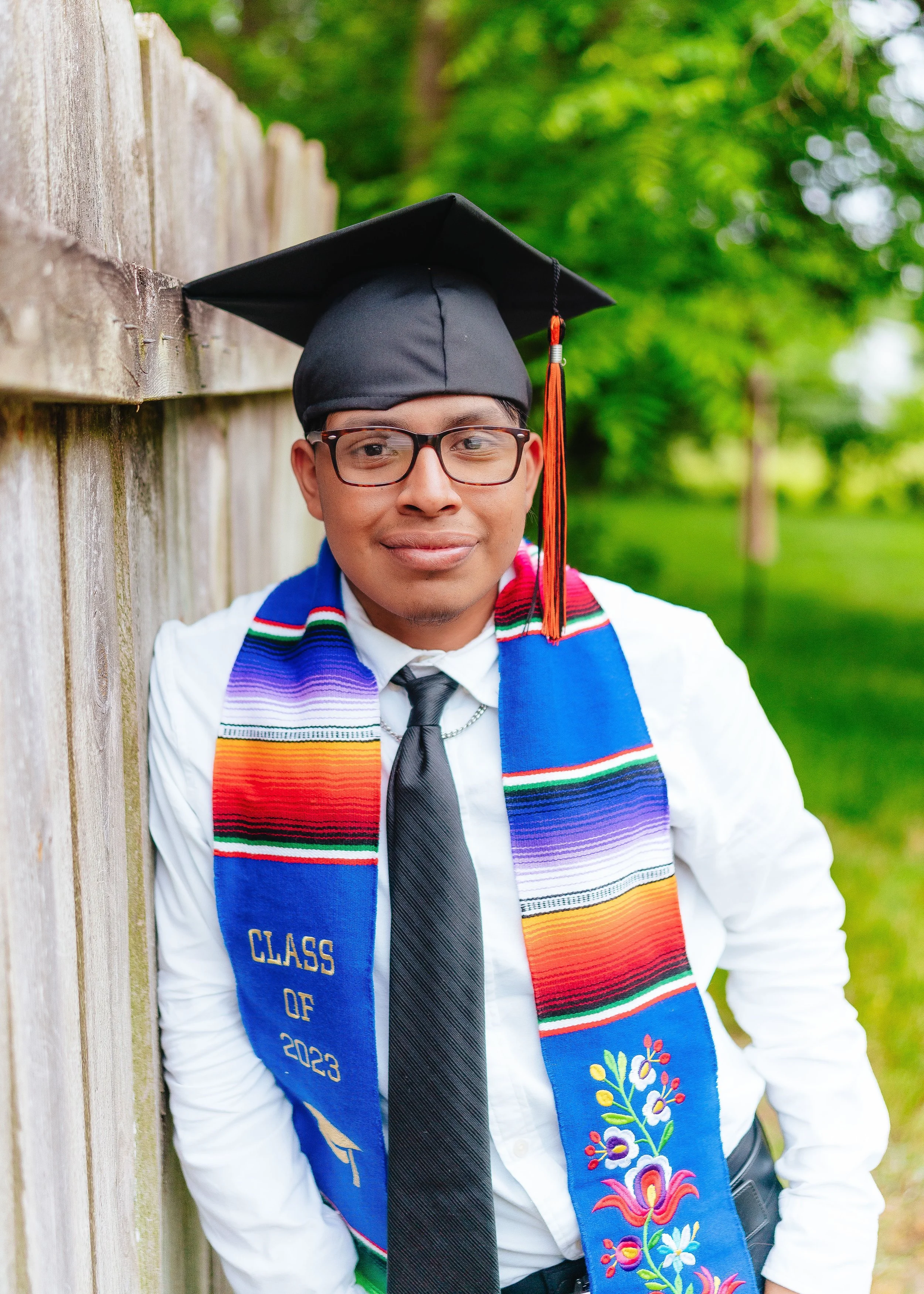 Young man in graduation cap and gown with a colorful stole, standing outdoors near a wooden fence, with green trees in the background.