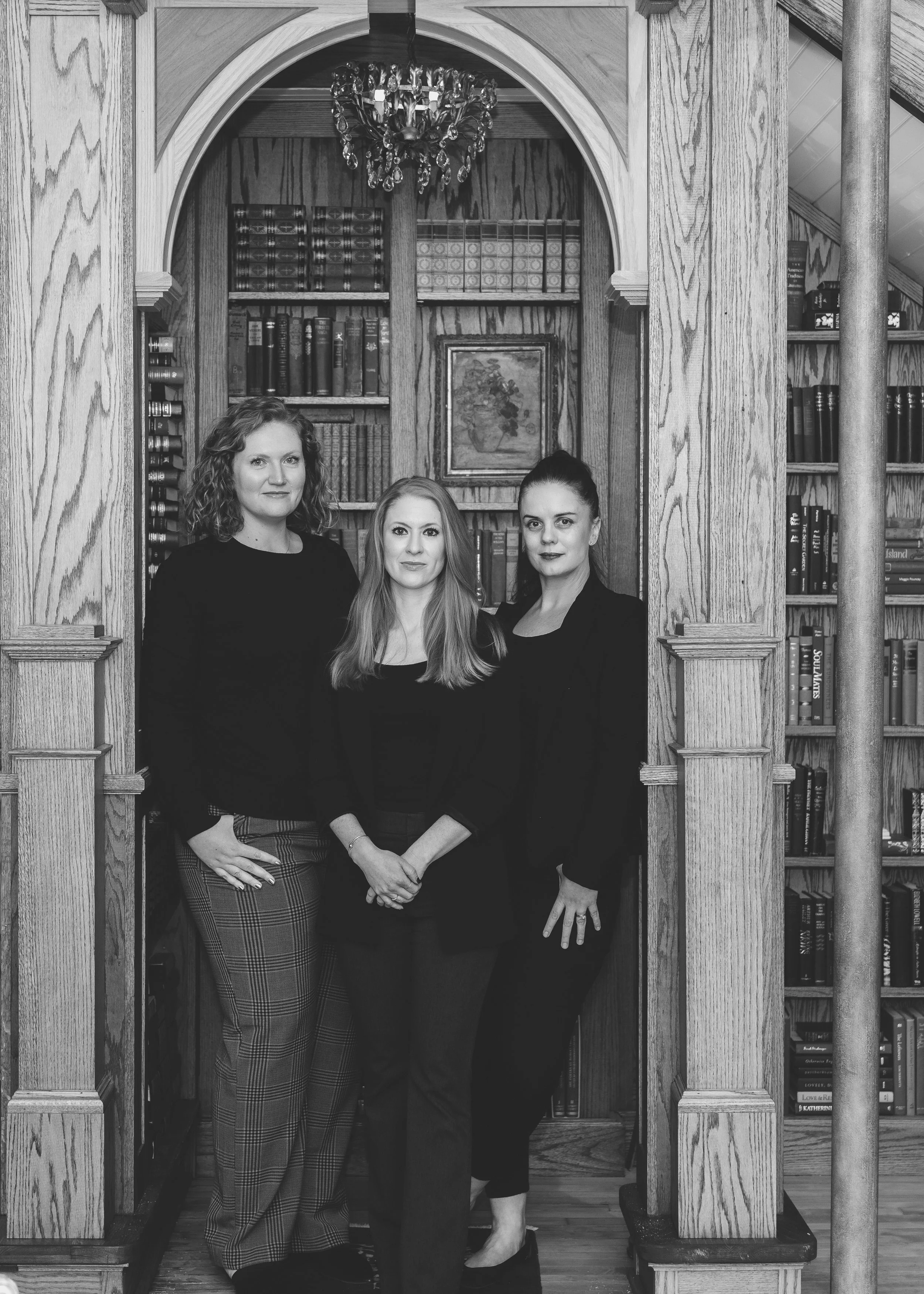 Three women standing together in front of wooden bookshelves filled with books, within a library or study room, with a chandelier hanging above.