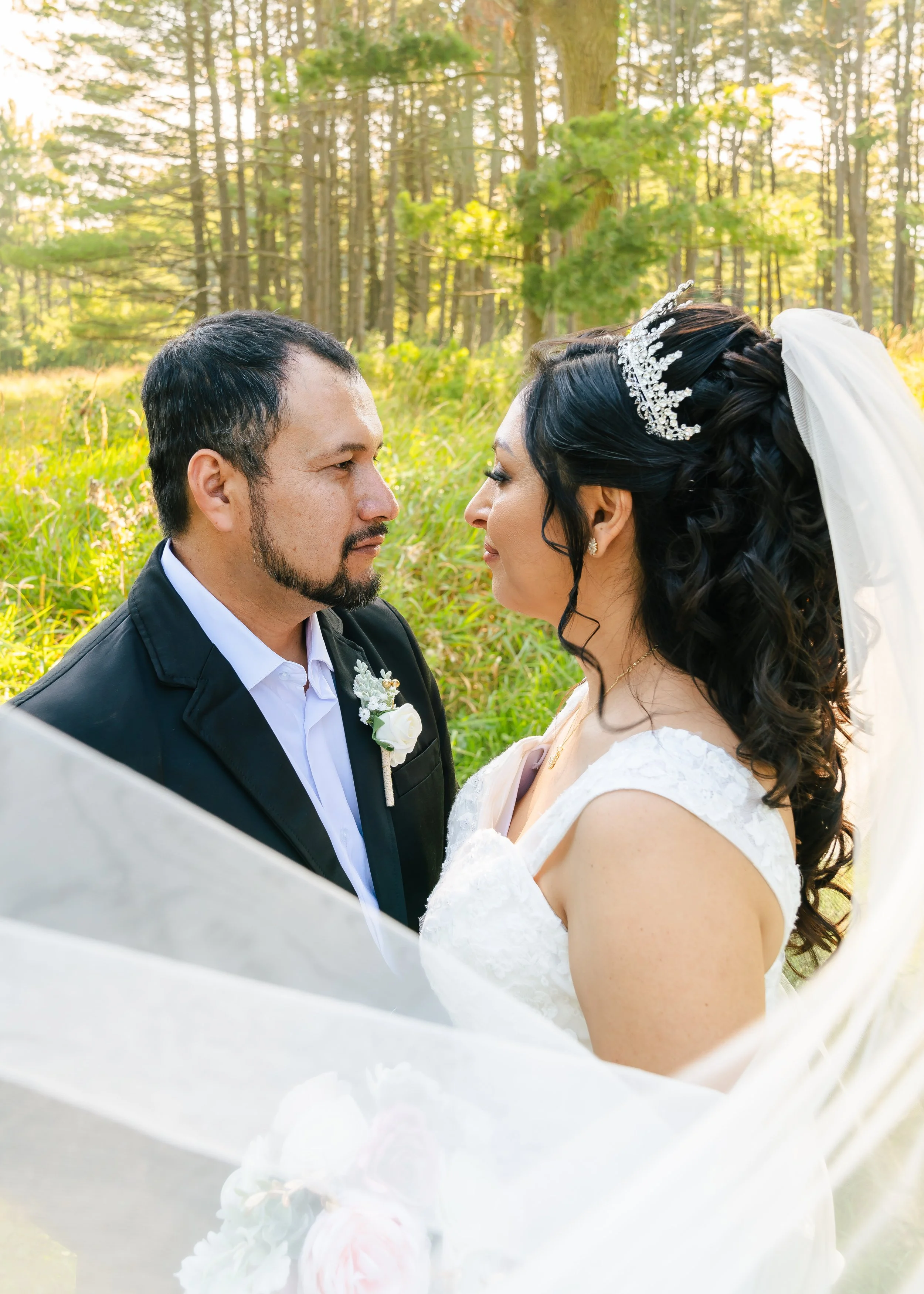 A bride and groom face each other in a forest during their wedding, with trees and sunlight in the background.