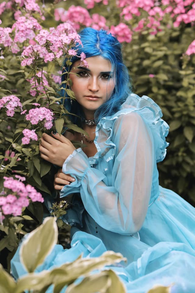 A woman with blue hair wearing a light blue dress, sitting among pink flowers and green foliage.