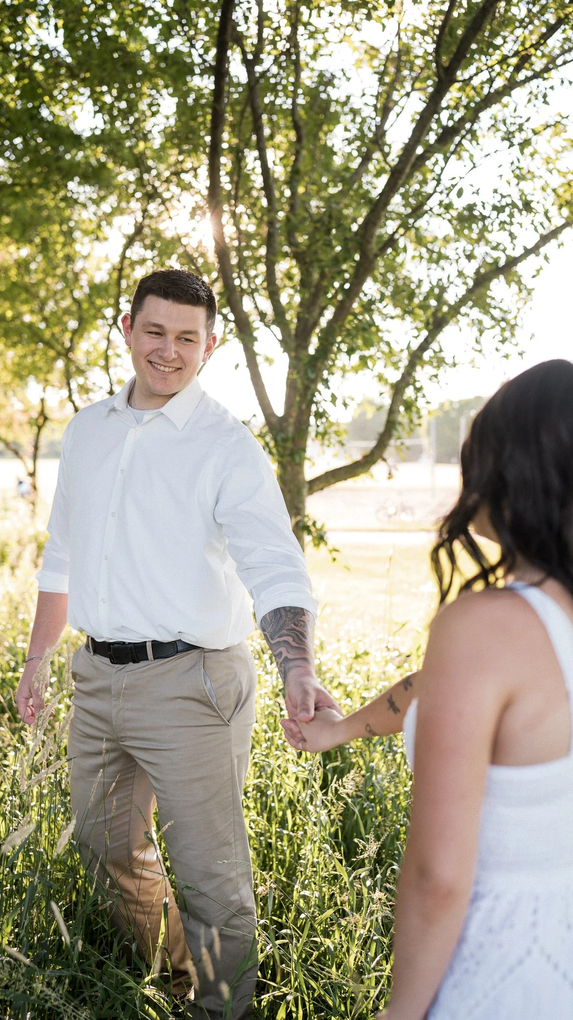 A young man in a white shirt and khaki pants smiling and holding hands with a woman in a white dress outdoors near a tree.