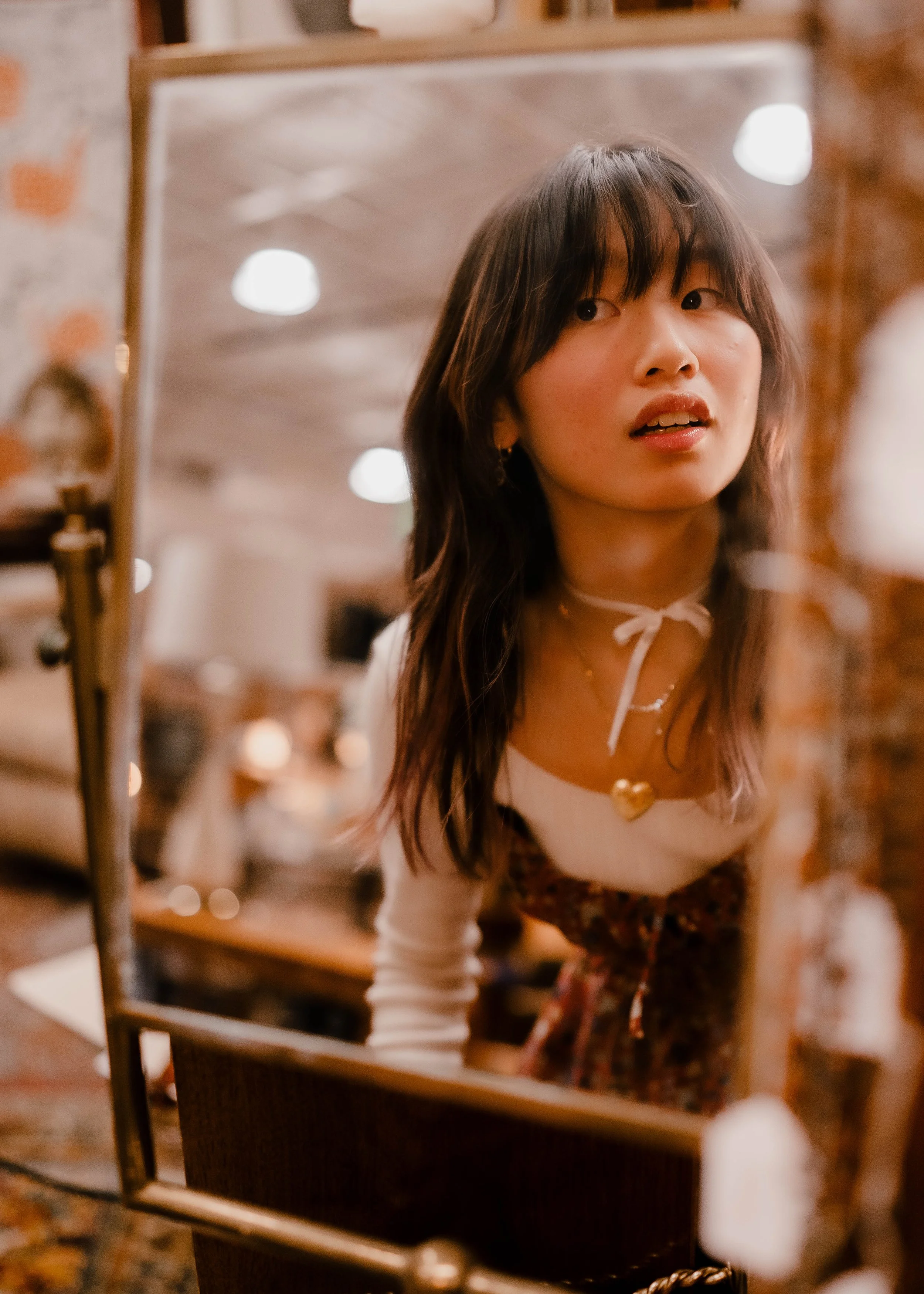 A young woman with shoulder-length dark hair looking into a mirror, with a blurred background of a cozy room, lit by warm lighting.