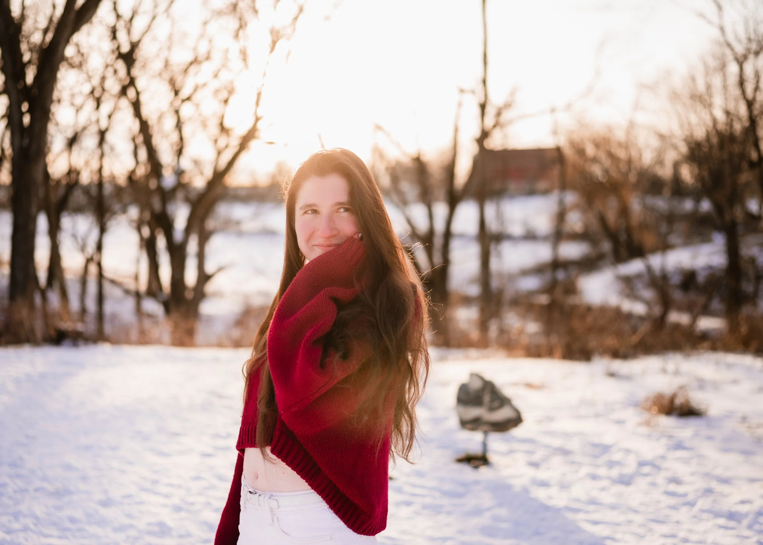 A young woman with long brown hair wearing a red sweater and white pants outdoors in a snowy landscape, smiling and looking back towards the camera with the sun setting behind her.
