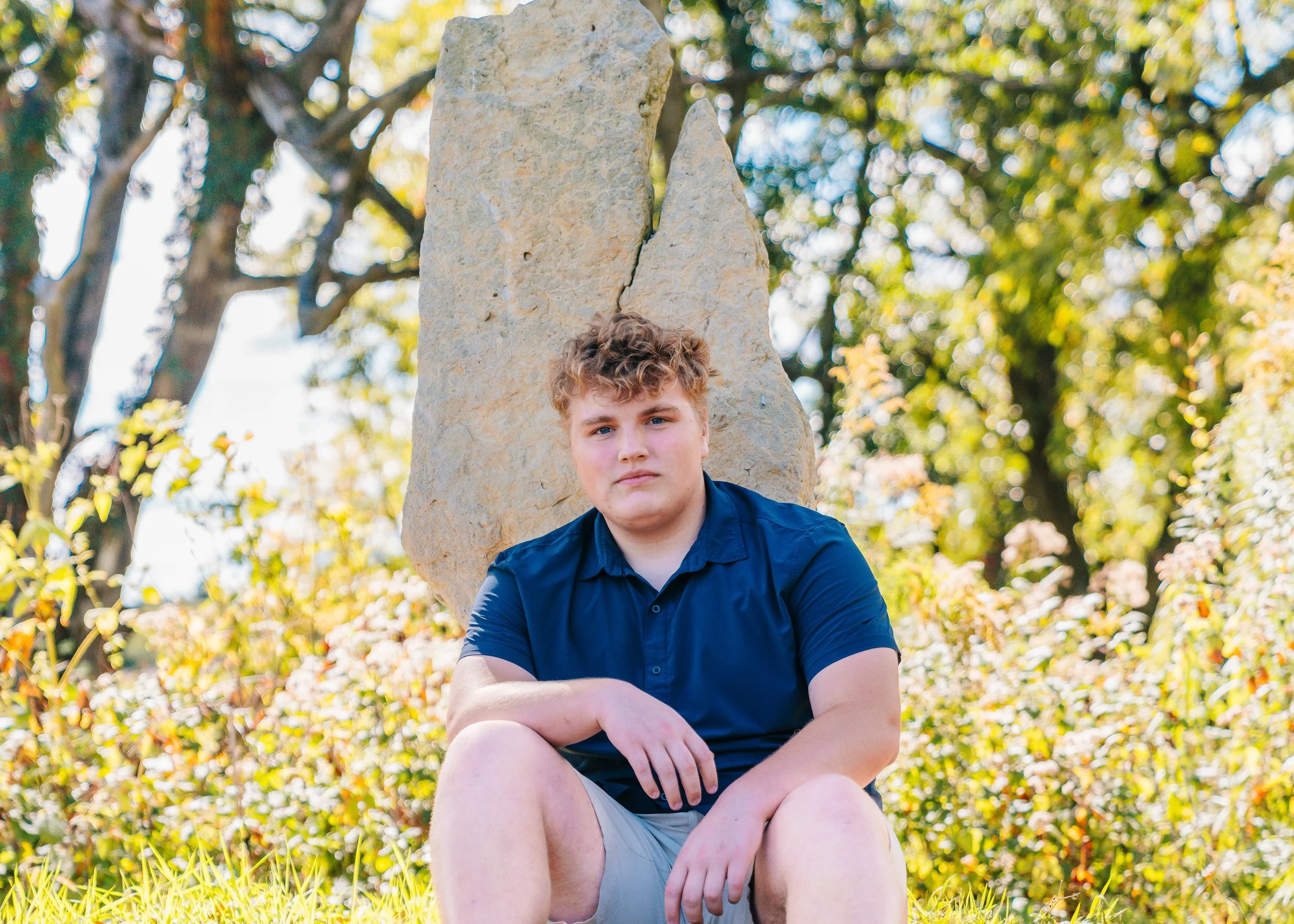 Young man with curly hair wearing a blue shirt and shorts, sitting on grass in front of a large tree and rocks in a sunny outdoor setting.