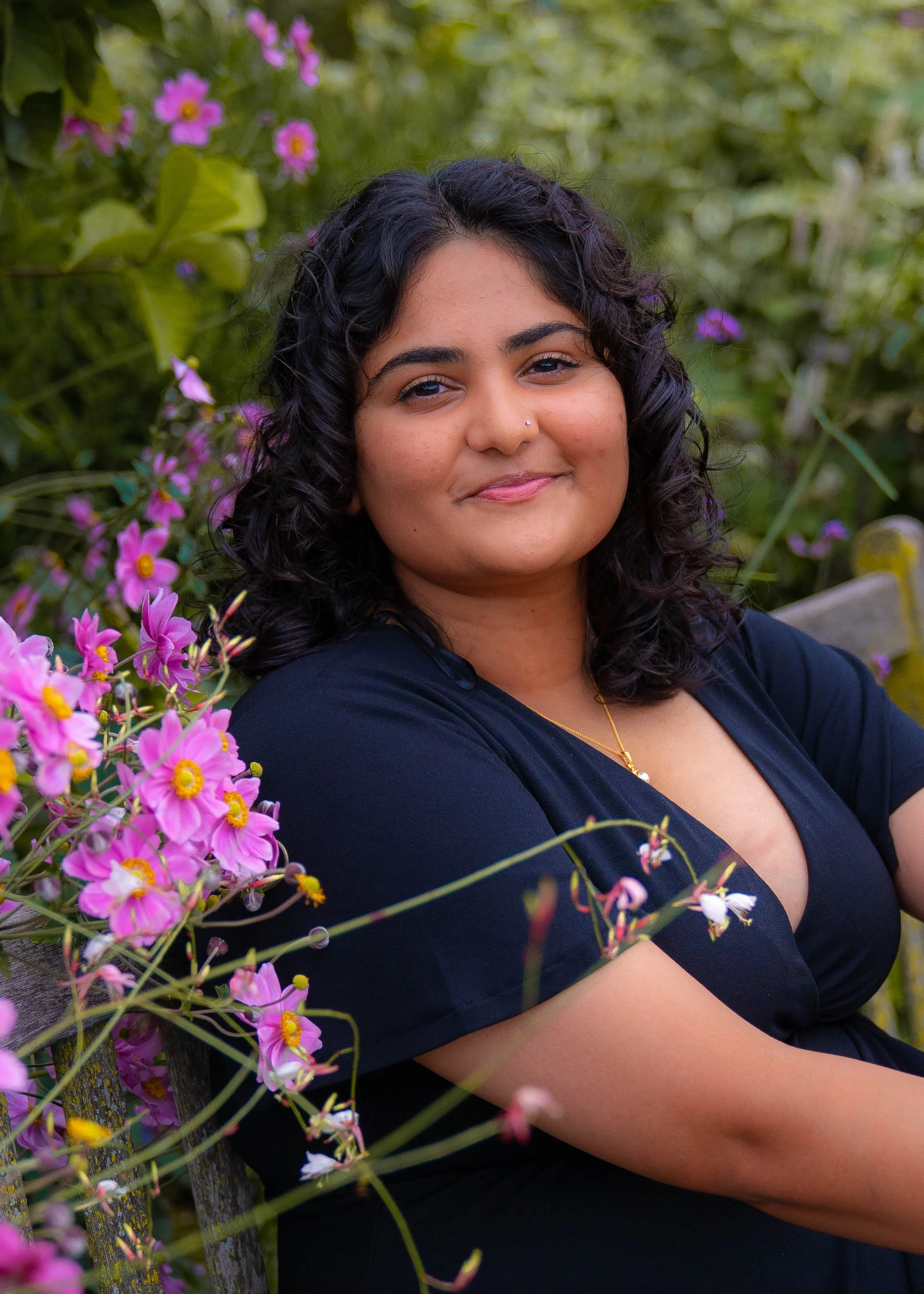 A woman with black curly hair and a nose piercing sits on a wooden bench surrounded by pink flowers outdoors, wearing a black top and a gold necklace.