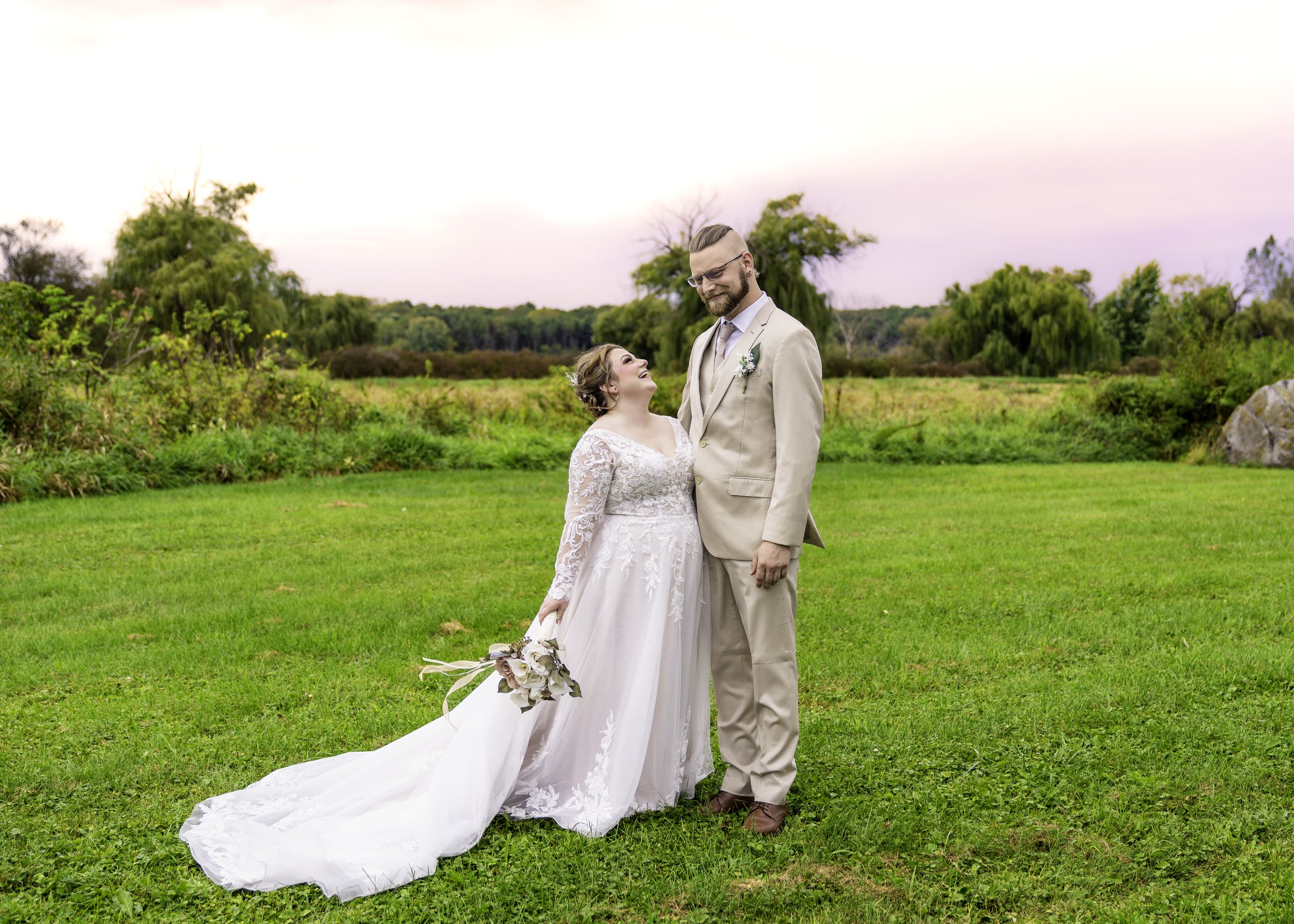 A bride in a white wedding gown holding a bouquet and a groom in a beige suit smiling at each other in a lush green field with trees in the background during sunset.