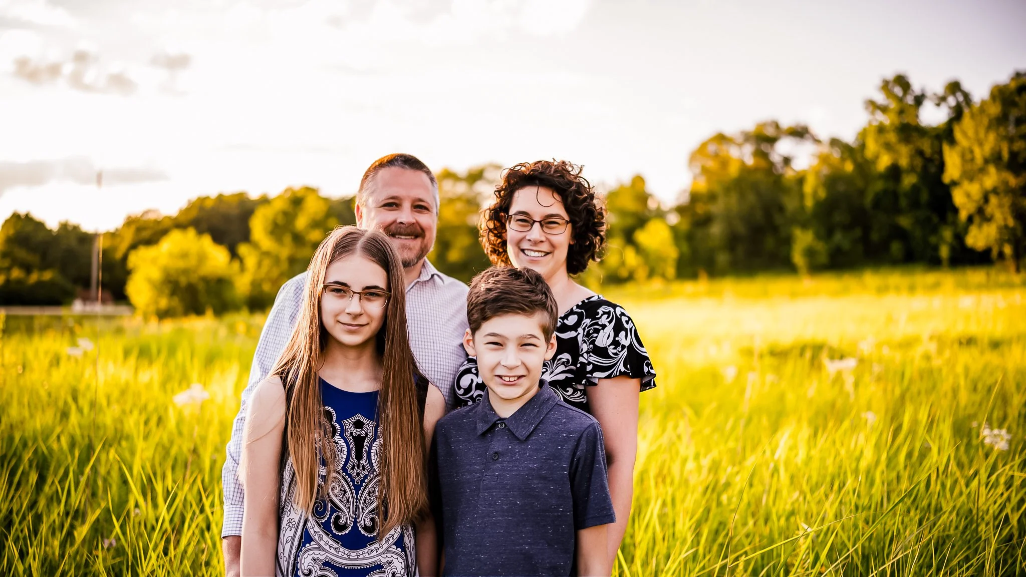 A family of four standing together in a grassy field during sunset, smiling at the camera. The family includes a man, a woman, a teenage girl, and a young boy, with trees in the background.