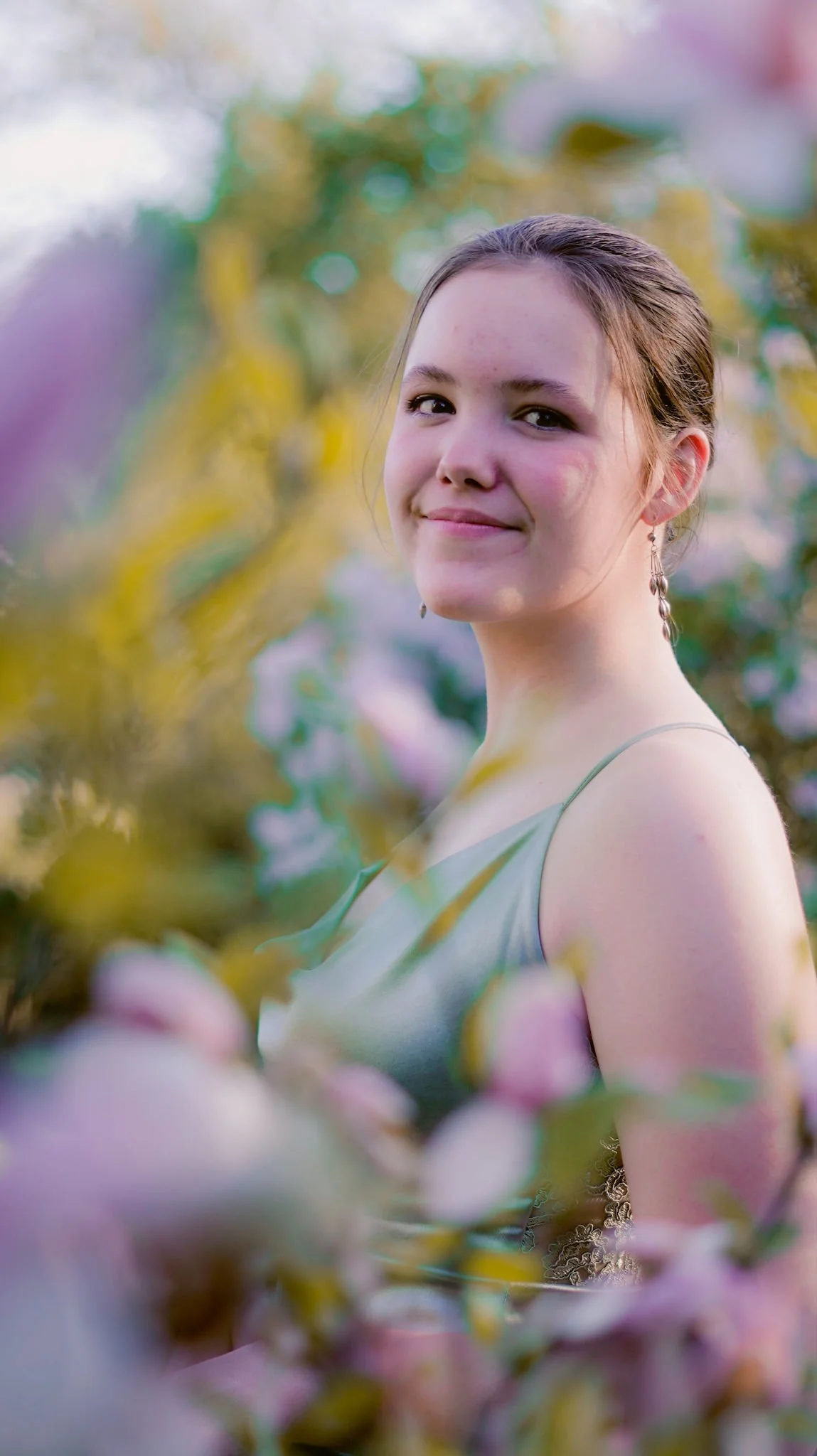 A young woman with a smile, wearing earrings and a light-colored dress, standing among pink blossoms outdoors.
