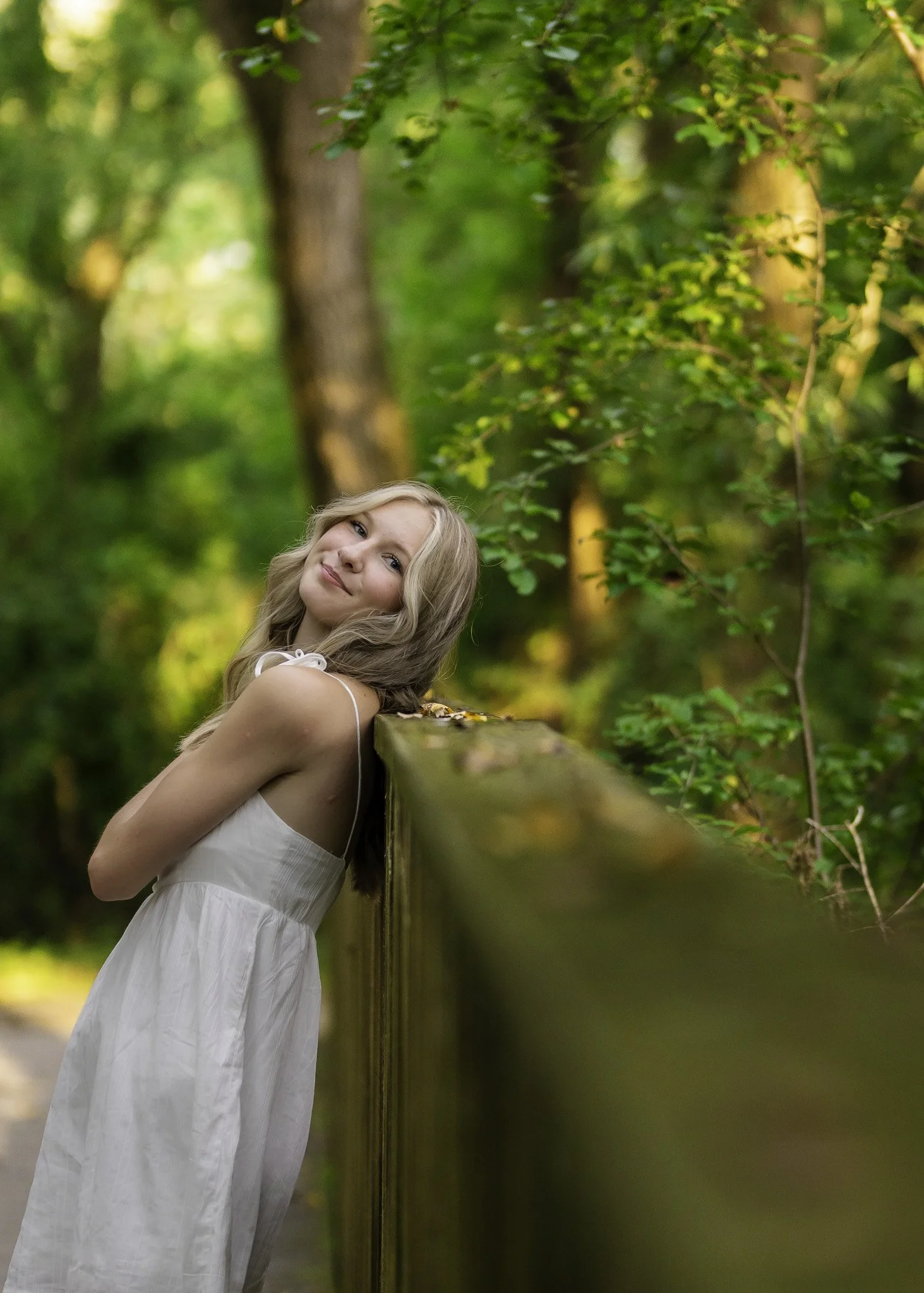 A young woman in a white dress leaning against a wooden fence in a forested area with lush green trees and dappled sunlight.